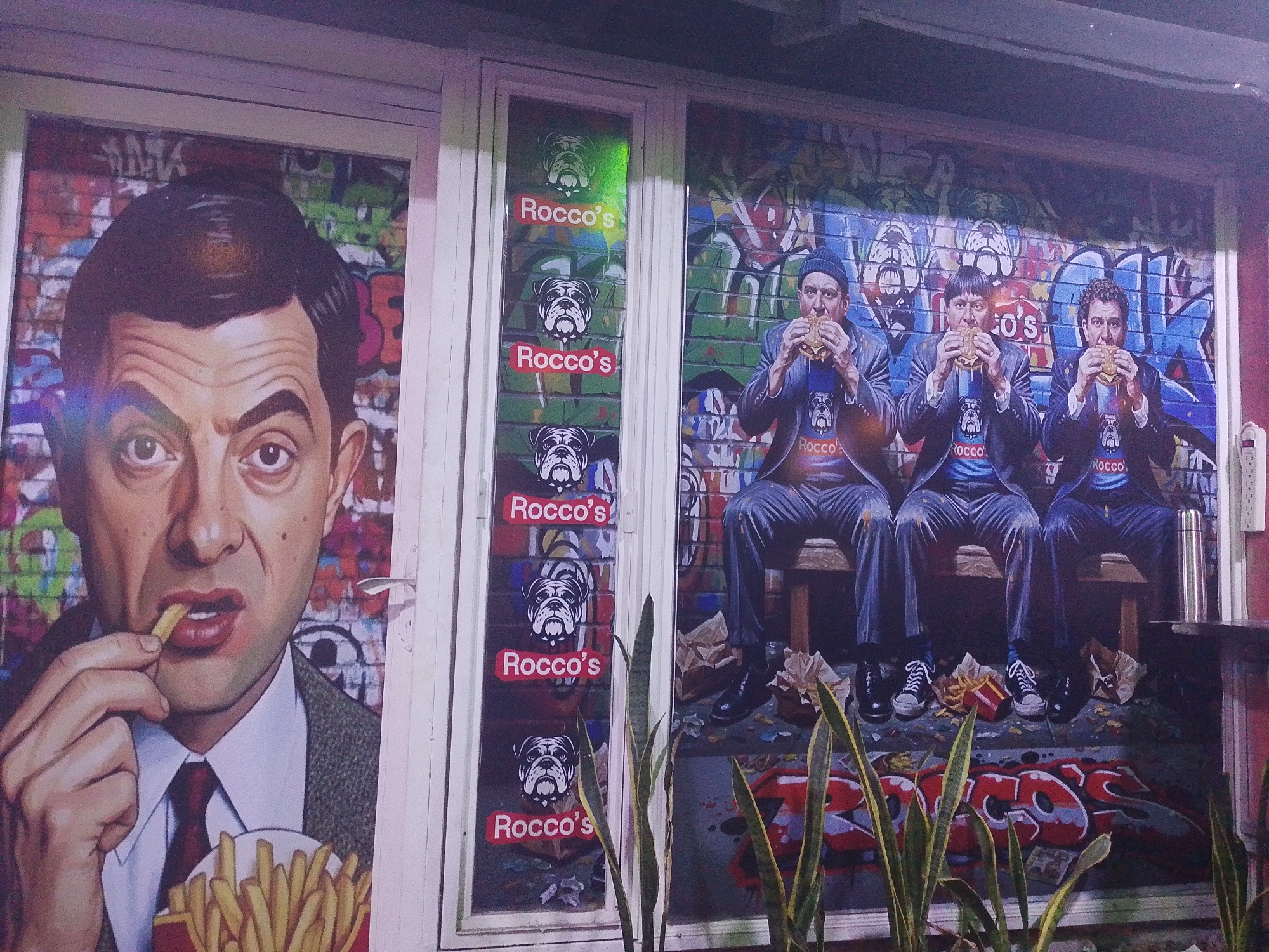 This is an outdoor scene in Tinaquillo, Venezuela. The exterior of a building, likely a restaurant, is shown. The building has multiple windows with murals painted on them. The left window features a large image of Rowan Atkinson's character, Mr. Bean, eating french fries. The middle window has the restaurant name "Rocco's" repeated multiple times with a bulldog head above each instance. The right window depicts three men resembling members of the Three Stooges, sitting on a bench and eating burgers. They are wearing suits and Rocco's t-shirts. The background is graffiti-style. "Rocco's" is also spray-painted at the bottom of the mural. In the foreground are tall, green plants. The scene appears to be at night or in low light.