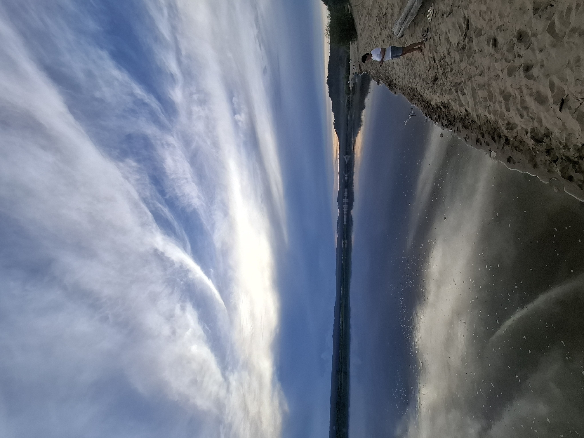 A person stands on a sandy riverbank, looking at their phone. The sky above is filled with wispy clouds, and the river reflects the sky and the distant treeline. The overall mood is serene and peaceful. The location is the city of Malaya Zazanka, Russia.