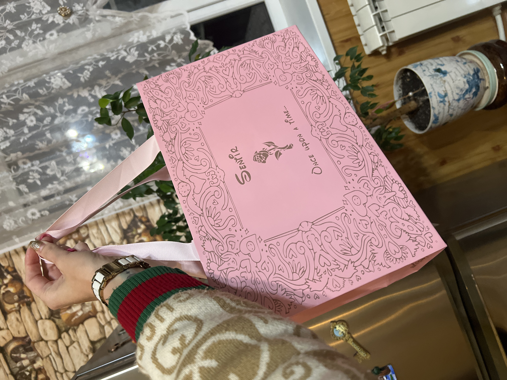 A person's hands are in the foreground, holding a pink gift bag with ornate brown line art. The bag is decorated with hearts, flowers, and whimsical figures. Text on the bag reads "ScentiR" and "Once upon a time...". The person is wearing a cream-colored sweater with a repeating pattern and a watch with a white and gold band. The background shows a lace curtain with floral patterns, a houseplant in a decorative blue and white pot, and wooden surfaces. The lighting suggests it might be daytime, possibly indoors with natural light filtering through the window. The overall mood conveyed is one of receiving or giving a special gift.