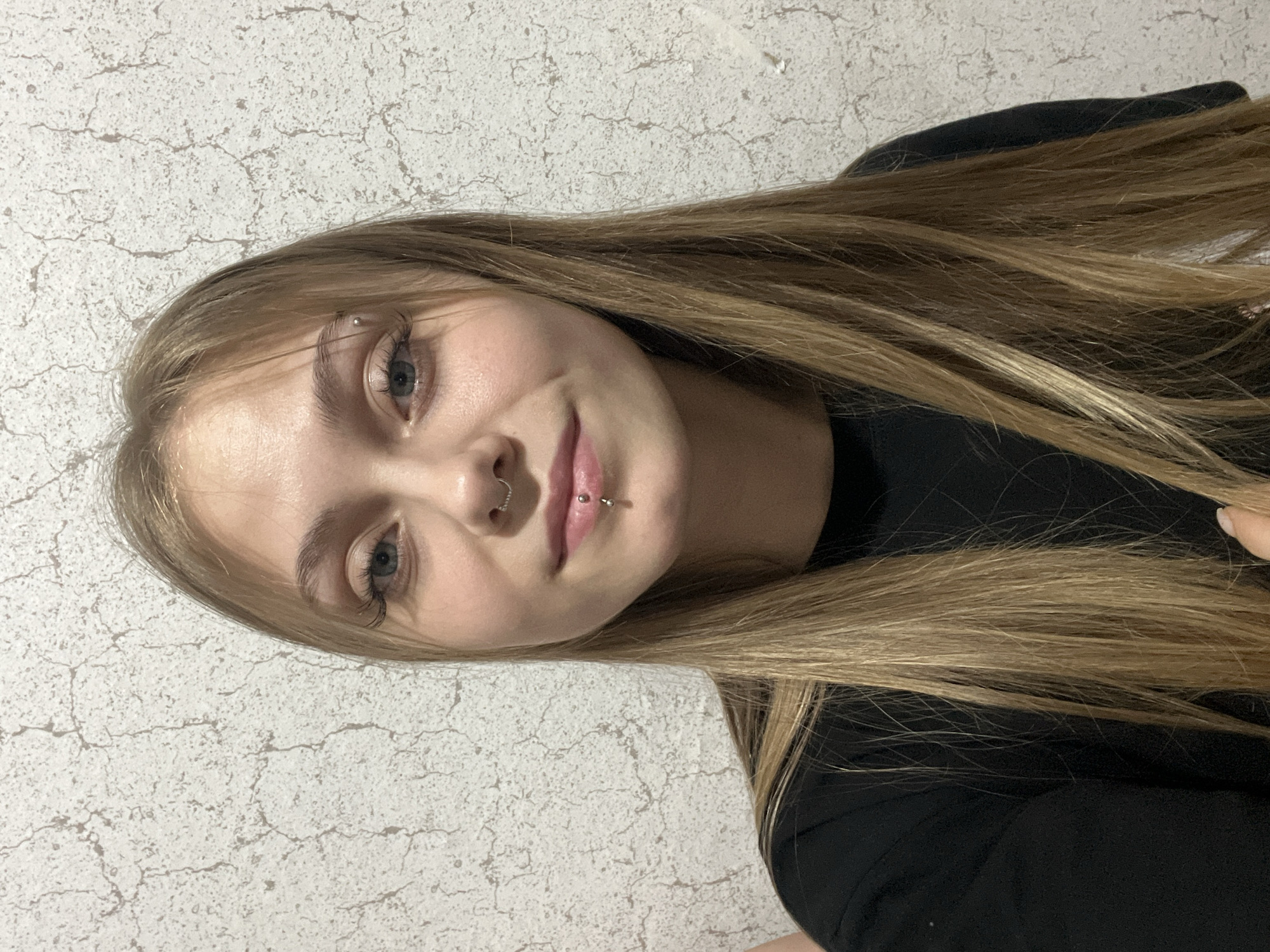 A young woman with long, straight, light brown hair and blue-grey eyes is shown in a rotated, close-up portrait. She has a calm expression with a subtle, closed-mouth smile, looking directly at the camera. Notable facial piercings include a decorative heart-shaped septum ring, a barbell-style philtrum piercing, and a small barbell on her right eyebrow. She is wearing a plain black top. The background is a textured wall, primarily white or off-white, with a pattern resembling cracks or distressed plaster in light brown or beige tones. The lighting suggests an indoor setting, possibly artificial. The image appears to be a posed photograph, potentially a selfie, taken in Фоки, Russia. No text is visible in the image.