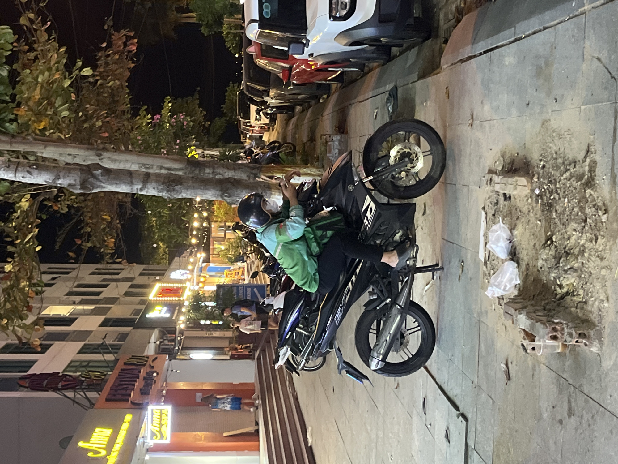 In the city of Bac Nha Trang Ward, Vietnam, a person wearing a green jacket, black pants, and a helmet sits on a black motorcycle on a sidewalk. The person is holding a smartphone and appears to be interacting with it. The motorcycle is parked next to a row of cars. The sidewalk is paved with gray tiles and shows signs of wear and tear, with some debris and a small pile of bricks. Behind the motorcycle, a building with multiple floors and illuminated windows is visible. Trees with dark leaves line the street, and string lights are visible in the distance, suggesting a commercial or entertainment area. The scene is captured at night, indicated by the artificial lighting.