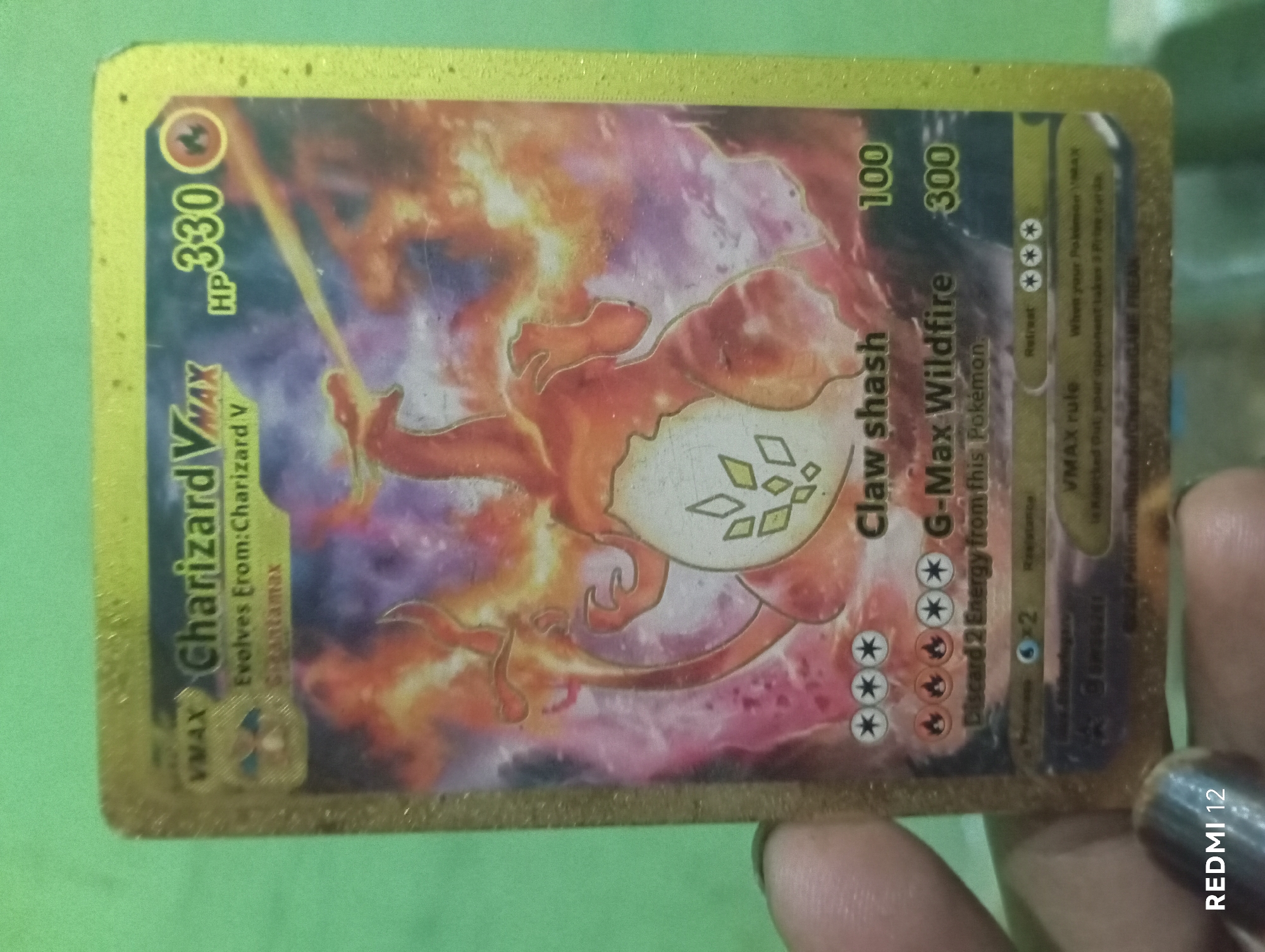 A close-up, eye-level, indoor shot shows a person's fingers holding a golden Pokémon trading card against a plain green background. The card features a fiery Charizard VMAX artwork with text and numbers indicating its game statistics. The card is slightly worn, with some smudges visible on its surface. The lower left corner of the image contains a watermark indicating "REDMI 12." The environment appears to be a simple room with no other discernible features or context suggesting a specific city or location.