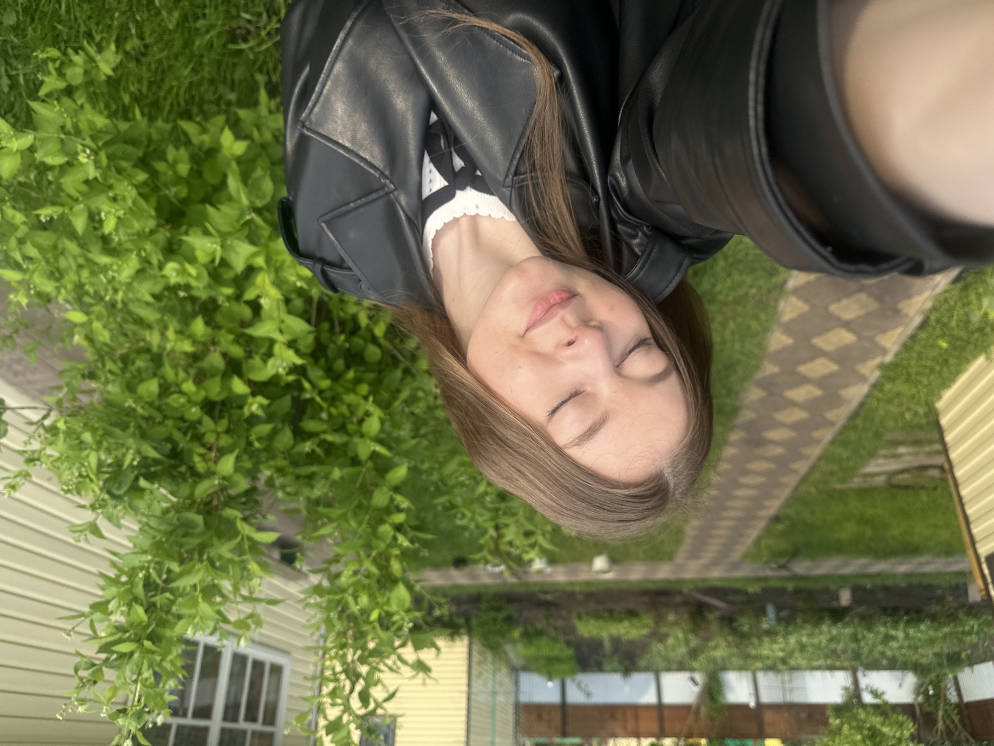 A selfie taken by a person with closed eyes, wearing a black leather jacket and a white sweater with a black pattern. The location appears to be outdoors in Коноваловка, Russia, based on the presence of vegetation, a building with a light-colored exterior, a paved walkway, and a garden or yard setting. The scene is well-lit, indicating daylight. There is greenery visible, suggesting a residential or suburban area.