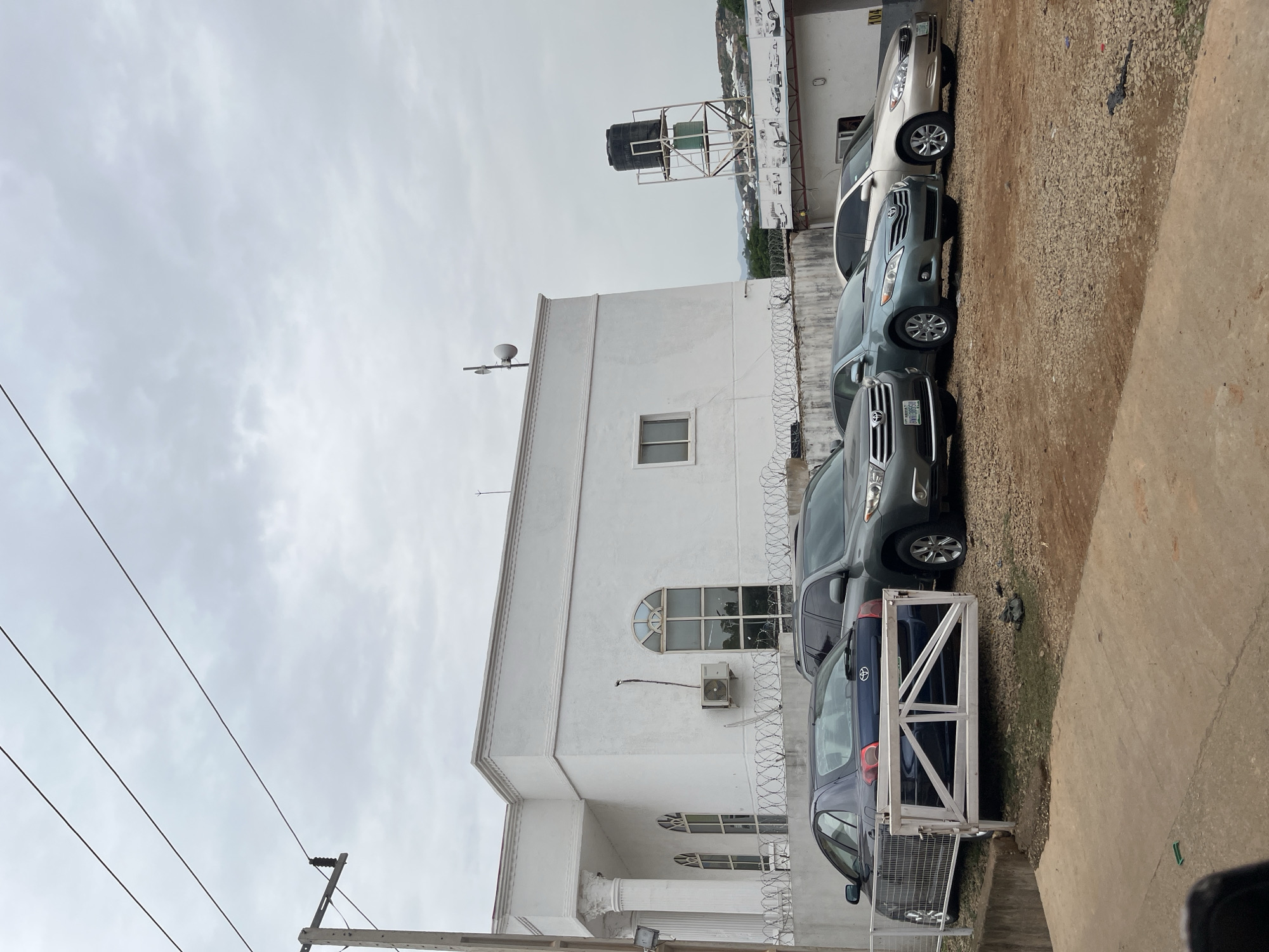 The image depicts a white building in Jos, Nigeria, with several cars parked in front of it. The building has a modern architectural style with columns and arched windows. A security fence with barbed wire runs along the front of the property. In the foreground, a dark blue sedan is partially visible behind a white metal gate. Behind it are several other vehicles, including a gray SUV and two sedans, one light blue and one gold. A water tank on a metal platform is visible on the roof of a smaller building to the right, and a speaker system is mounted on the roof of the main building. The sky is overcast, suggesting a cloudy day. The ground in the parking area is covered with gravel.