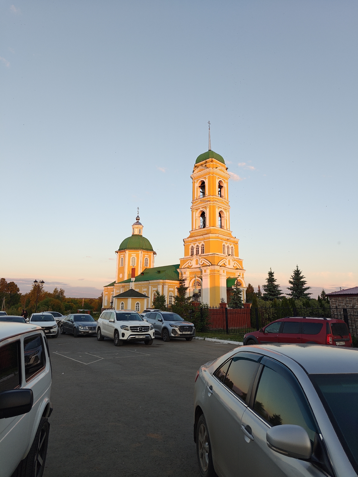 The image depicts the St. Nicholas Church (Nikolsky Cathedral) in Nikolo-Beryozovka, Russia, captured during late afternoon or early evening. The prominent church features a vibrant yellow facade with white decorative trim and contrasting green domes and roofs. A tall, multi-tiered bell tower, also yellow and green, dominates the right side of the structure, topped with a spire and cross. A smaller dome with a cross is visible on the main church building to the left of the bell tower.
In the foreground, an asphalt parking lot is present, occupied by several vehicles. Partial views of a white SUV (resembling a Lada Niva) are visible on the far left, and a silver sedan (resembling a Toyota Camry) is partially visible on the far right. In the middle ground, parked cars include a white Mercedes-Benz SUV, a dark gray sedan, a white sedan, a silver-gray SUV, and a red crossover. Faded white lines mark parking spaces on the asphalt.
A dark metal fence with red brick pillars separates the parking area from the church grounds, where several dark green conifer trees are planted near the fence line. Deciduous trees with some autumn foliage are visible in the background, further from the church. The sky is a gradient of light blue at the top, transitioning to warm orange and pink hues near the horizon, indicative of sunset. Numerous small, dark specks, likely birds or insects, are scattered across the sky. The low sun casts strong, warm light on the church's upper sections, creating highlights and shadows. A few indistinct human figures are visible near cars in the mid-ground on the left side of the image.
