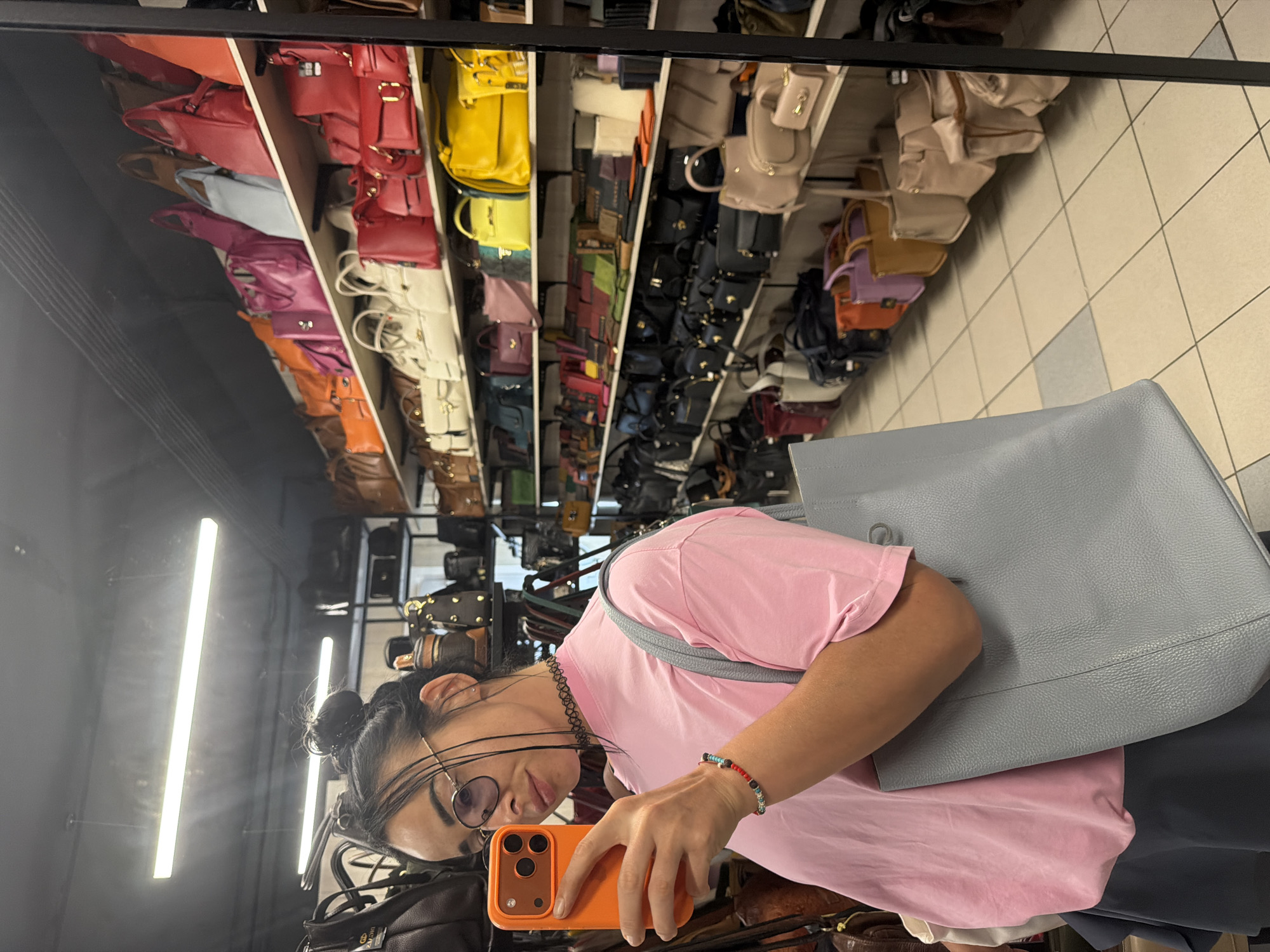 A woman with dark hair pulled into a bun, wearing glasses, a pink t-shirt, and a grey skirt, takes a mirror selfie. She is holding an orange smartphone in her right hand and a large grey tote bag over her left shoulder. She is wearing a black choker necklace and a beaded bracelet on her left wrist. The background shows shelves stocked with numerous handbags of various colors and styles. This scene appears to be inside a retail store. This image was taken in Sochi, Russia.
