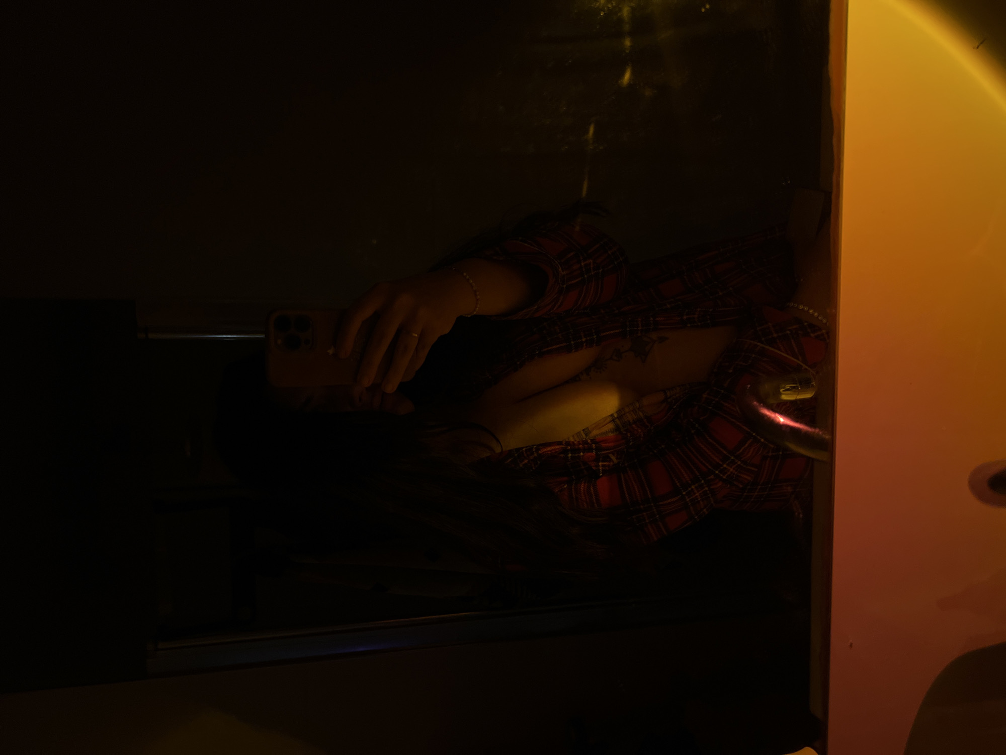 A woman is taking a selfie in a darkened room. She is wearing a red and black plaid shirt. She is holding a smartphone in her left hand. Her face is partially obscured by the phone and shadow. There is a tattoo visible on her chest. To her right is a wall that is lit with orange light. The location in the city of Kiryat Bialik, Israel is not discernible from the image.