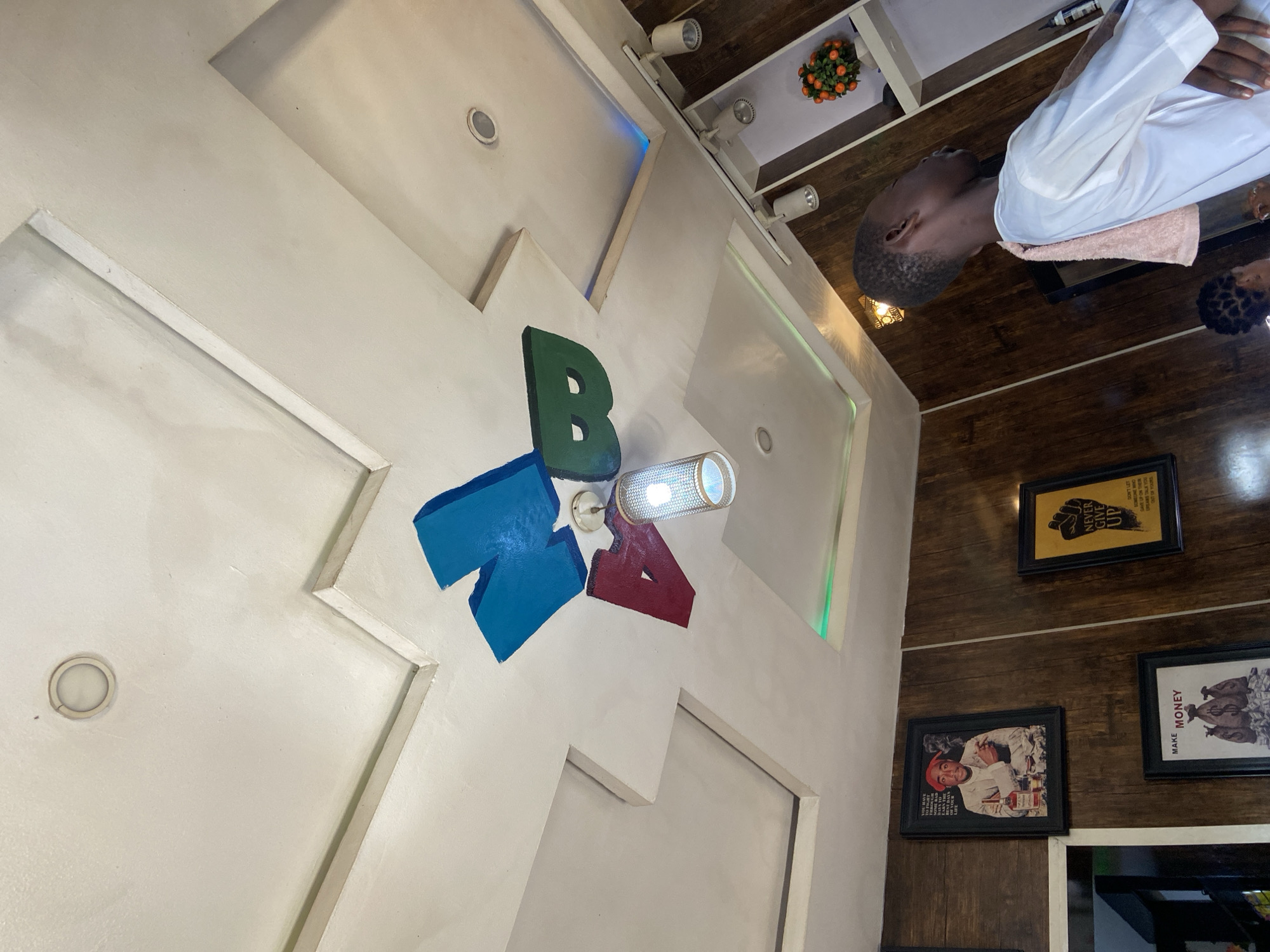 The image shows the interior of a building, possibly a business or establishment in Lokoja, Nigeria. A man in a white shirt is sitting at a table. The walls are decorated with framed pictures and a logo of the letter "BRA" in green, blue, and red. The ceiling has recessed lighting and a decorative arrangement of oranges. The walls are a light color.
