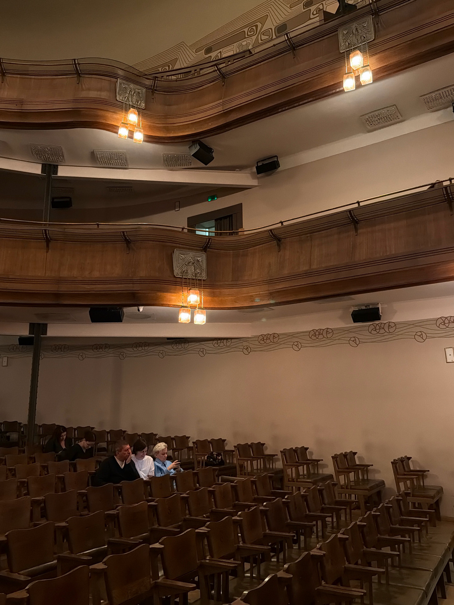 The image depicts the interior of a theater with tiered seating. In the foreground, rows of empty wooden chairs are visible, some with dark green or brown upholstered seats. Further back, a few people are seated, including a man in a dark sweater and a woman in a white shirt, both looking forward. Another woman in a blue shirt is looking at her phone. The theater features ornate wooden balconies with decorative metal railings. Hanging from the upper balcony are several square, amber-colored glass light fixtures. The walls are a light beige color, with decorative patterns in a darker color along the upper sections. The lighting is warm and dim, creating an intimate atmosphere. This appears to be the interior of the Meyerhold Theater in Moscow, Russia.