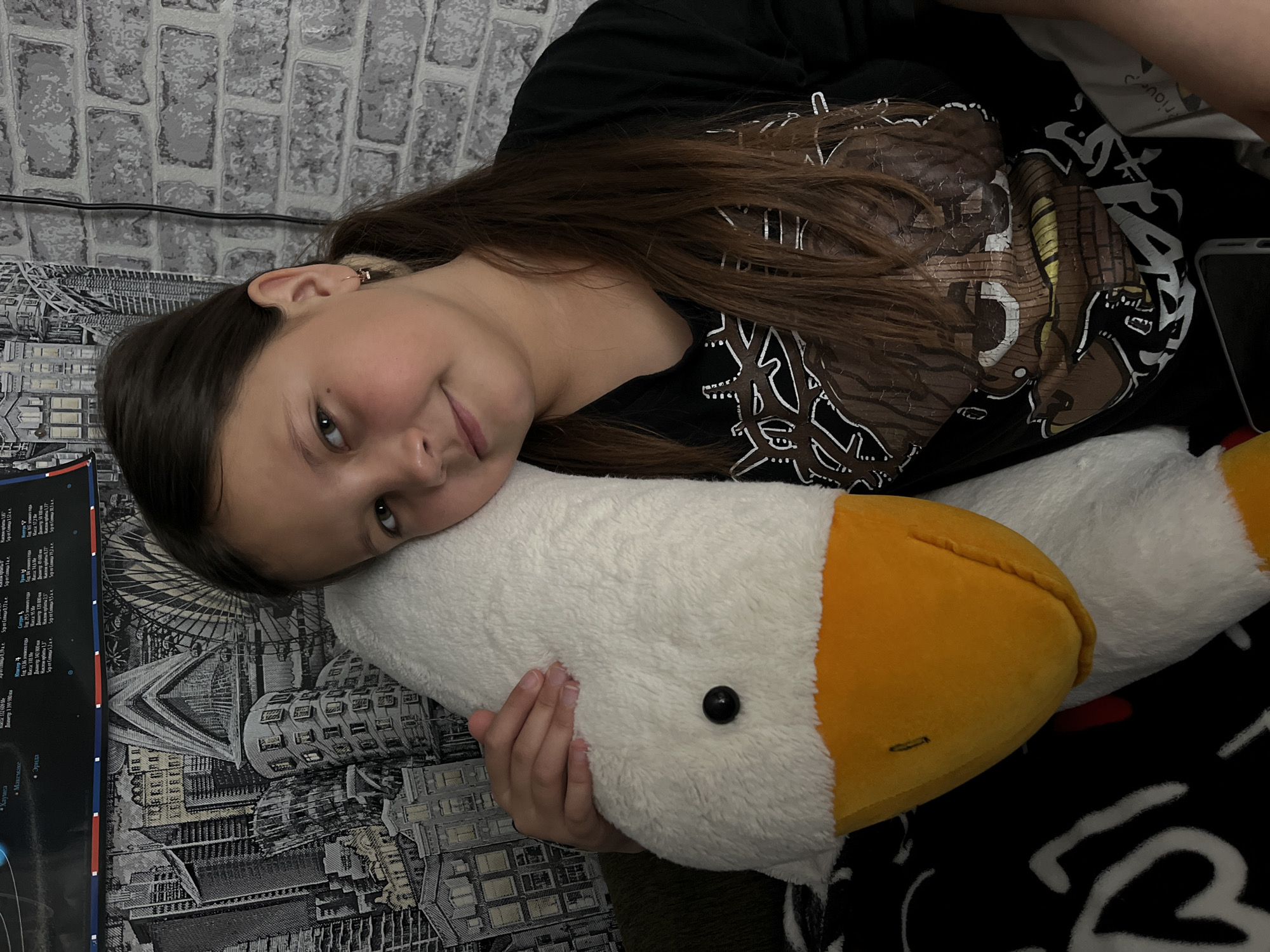 A girl with long brown hair lies on her side, resting her head on a large, stuffed white goose with a yellow beak. She is wearing a black t-shirt with a dark design. The background consists of a gray brick-patterned wall and a textured blanket depicting a cityscape. To the left of the girl is a poster with text in a Cyrillic alphabet. The image appears to be taken indoors.