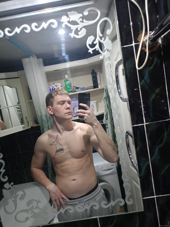 A male individual is depicted taking a mirror selfie in a bathroom located in Chaikovsky, Russia. The male, appearing to be in his late teens or twenties with short, light-brown hair, stands shirtless, wearing grey sweatpants with a visible black underwear waistband. A black tattoo, depicting an animal head with two horizontal stripes across its neck, is visible on his left pectoral muscle. He holds a dark-colored smartphone in his right hand, positioned to capture his reflection, while his left hand rests on his hip.
The bathroom features dark green/black glossy wall tiles with white grout lines. The rectangular mirror has frosted floral and scrollwork designs along its upper, lower, and right edges. Reflected in the mirror, behind the individual, is a white front-loading washing machine. Above the washing machine, a light-colored open shelving unit holds various personal care products, including bottles of shampoo, conditioner, mouthwash, and other containers. To the left, a white shower curtain is partially drawn, revealing a glimpse of a white cabinet below. A chrome heated towel rail is visible to the right, and a light-colored towel is draped over it. A light source is reflected in the upper center of the mirror, and a light blue pipe is visible near the upper left corner of the reflection. Exposed wiring or a fixture with a brown base is observed in the upper right.