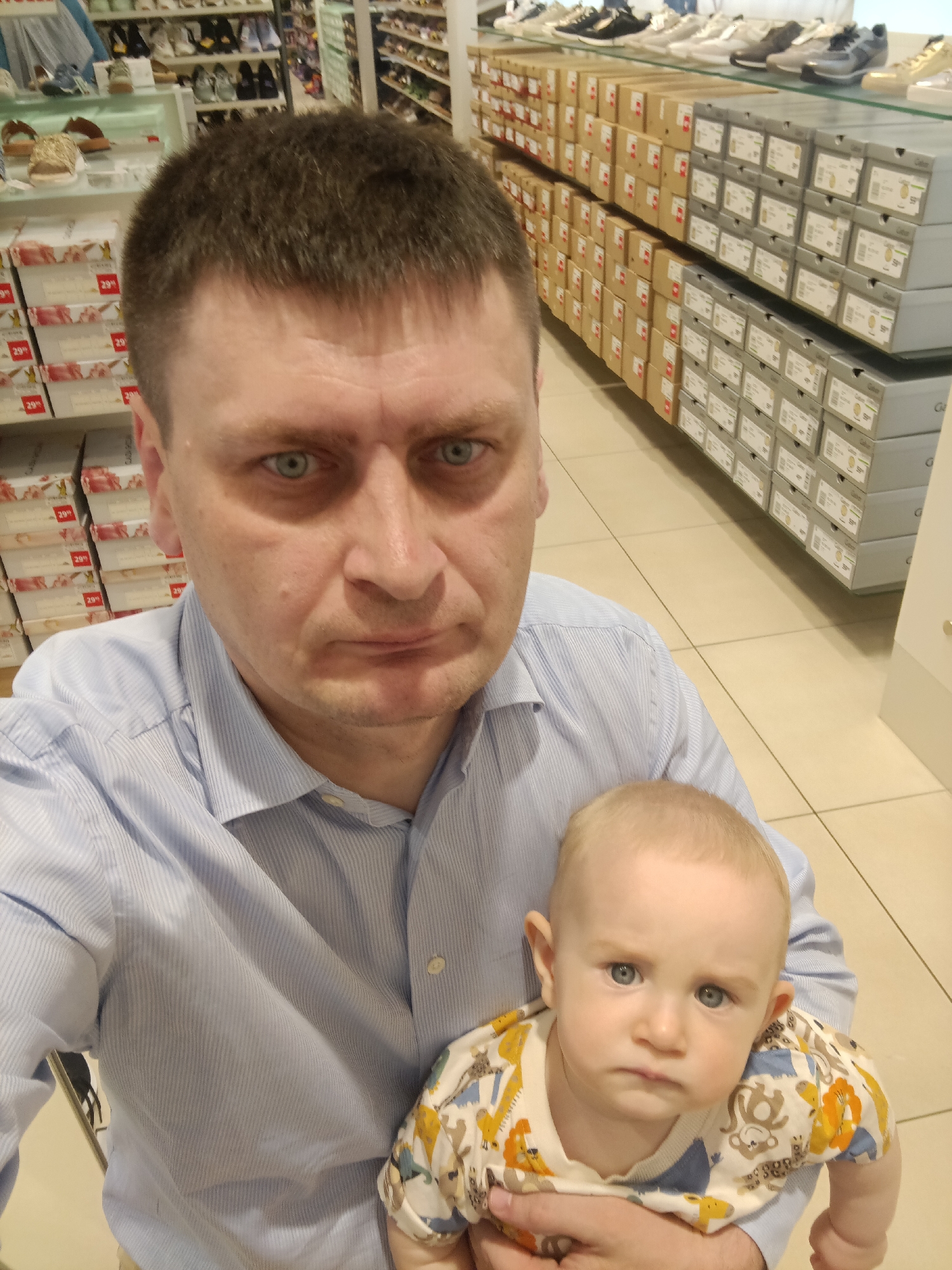 This media file shows a close-up selfie of a man holding a baby. The man, with short brown hair and blue eyes, wears a light blue striped button-down shirt. He is looking directly at the camera with a somewhat serious expression. The baby, held in the man's arms, has fair skin, light hair, and blue eyes. The baby is wearing a light-colored onesie with a repeating pattern of cartoon animals like lions and monkeys.
The background suggests an indoor retail environment, likely a shoe store. Shelves filled with shoe boxes are visible in the background. Some boxes are stacked high, while others are neatly arranged on display shelves. The flooring appears to be tiled. The lighting is bright, consistent with an indoor retail space.
The man and baby are the primary subjects. They are positioned in the foreground, with the man's head and shoulders taking up a significant portion of the frame. The baby is nestled in the man's arms, facing slightly away from the camera.
The activity appears to be a casual moment captured by the man himself, possibly while shopping with his child. There are no explicit indications of a specific event or time of day, other than it being indoors.
On the shoe boxes, some visible text includes "29%" and what appears to be brand names or model numbers, though these are largely illegible. The setting clearly indicates a commercial establishment, likely a shoe store, given the abundance of shoe boxes. The emotional tone seems neutral to slightly serious for the man, and mildly curious or contemplative for the baby. The location context provided is Nuremberg, Germany, which adds a geographical element to the scene.