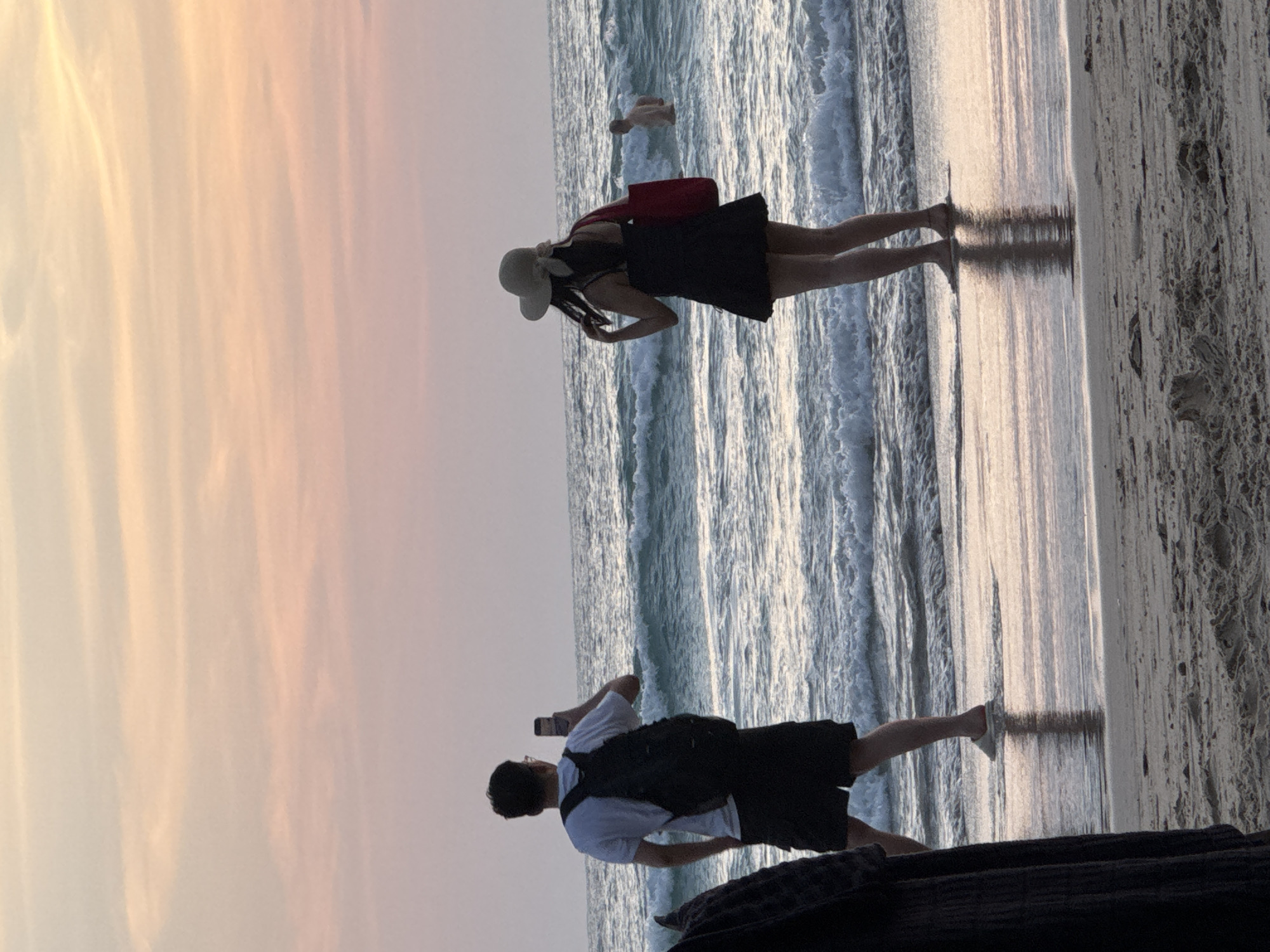 This image depicts a serene sunset scene at Karon Beach, Thailand. In the foreground, a man stands facing the ocean, holding a smartphone and appearing to take a photo. He wears a white t-shirt, black shorts, and a backpack. To his right, a woman is also facing the sea, wearing a black dress, a straw hat, and carrying a red bag. She is touching her hair. Further out in the ocean, a person is visible wading in the water. The gentle waves of the Andaman Sea are rolling onto the sandy shore, reflecting the soft hues of the pastel-colored sky, a mixture of pink and orange. The sand on the beach appears textured, with some small mounds and footprints. A dark, draped object is partially visible on the left side of the frame, adding a sense of depth.