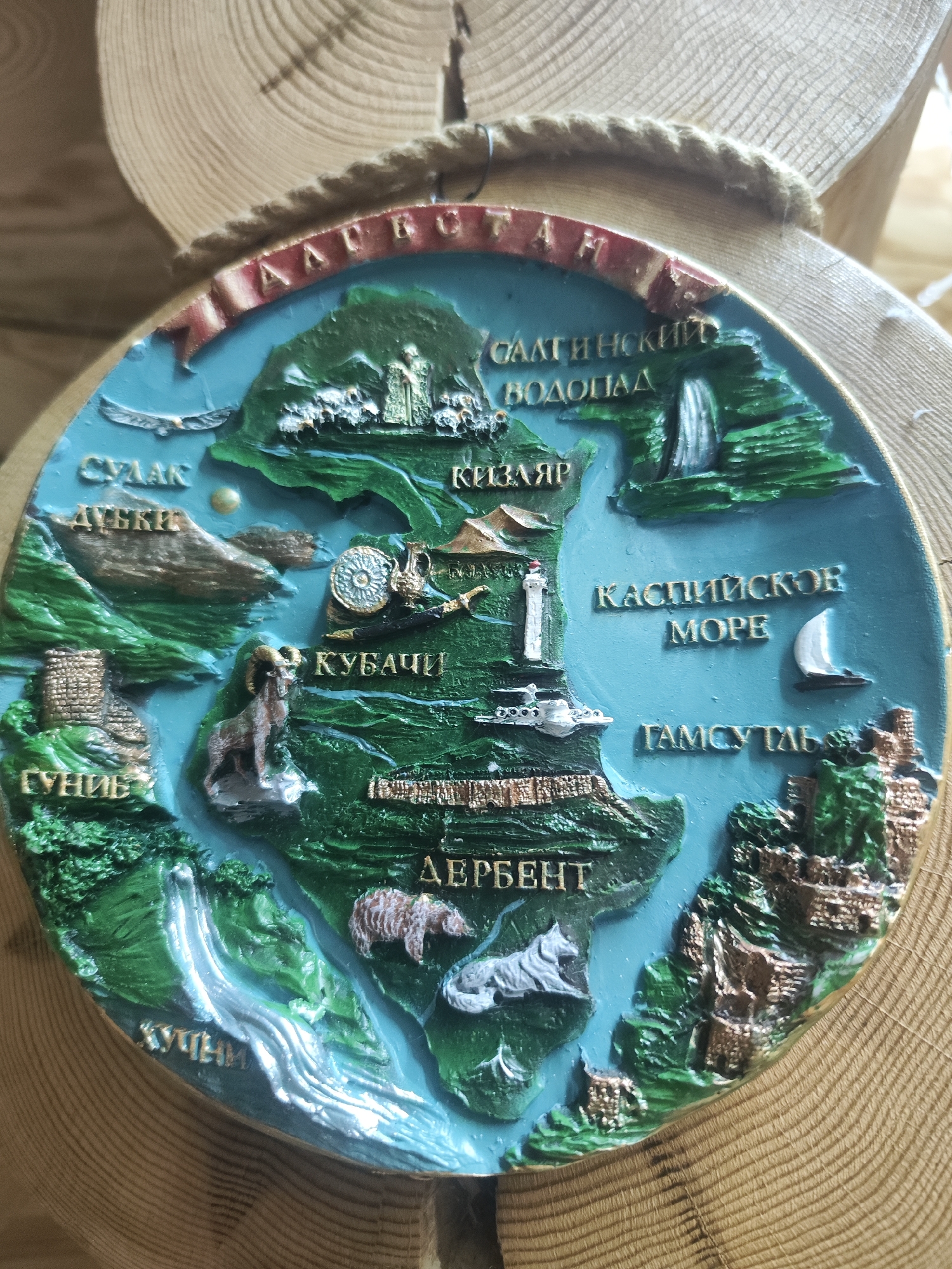 The image displays a round, relief-sculpted souvenir plaque or magnet, approximately 15-20 cm in diameter, resting against a light-colored wooden surface. The plaque vividly depicts a stylized map of the Dagestan region, showcasing its key geographical features, landmarks, and cultural symbols.
The landscape features prominent green landmasses, representing mountains and valleys, crisscrossed by blue rivers. A large blue body of water, labeled "КАСПИЙСКОЕ МОРЕ" (Caspian Sea), dominates the right side and includes a small white sailboat.
Visible text and associated elements in Cyrillic script include:
* A red banner across the top reads "ДАГЕСТАН" (DAGHESTAN).
* In the upper left, "СУЛАК" (Sulak) is written, with an eagle flying above.
* Below Sulak, "ДУБКИ" (Dubki) is situated next to a mountain.
* In the upper right, "САЛТИНСКИЙ ВОДОПАД" (Saltinsky Waterfall) is depicted as a cascading waterfall.
* Below the waterfall, "КИЗЛЯР" (Kizlyar) is marked.
* Mid-left, "КУБАЧИ" (Kubachi) is accompanied by an ornate disk, a jug, and a saber, alongside a statue of a mountain goat/ram.
* A white lighthouse stands near the Caspian Sea.
* Mid-right, "ГАМСУТЛЬ" (Gamsutl) is illustrated as an ancient, multi-tiered mountain settlement.
* In the lower left, "ГУНИБ" (Gunib) is represented by a fortified structure.
* Along a river in the bottom-left, "ХУЧНИ" (Khuchni) is visible.
* At the bottom center, "ДЕРБЕНТ" (Derbent) is shown with fortifications and two animal figures, a bear and a wolf-like creature.
The plaque is rich in detail and color, highlighting the natural beauty and historical significance of Dagestan. It appears to be a commemorative or decorative item, likely intended for tourists. The background consists of a wooden log or cross-section, showing distinct growth rings.