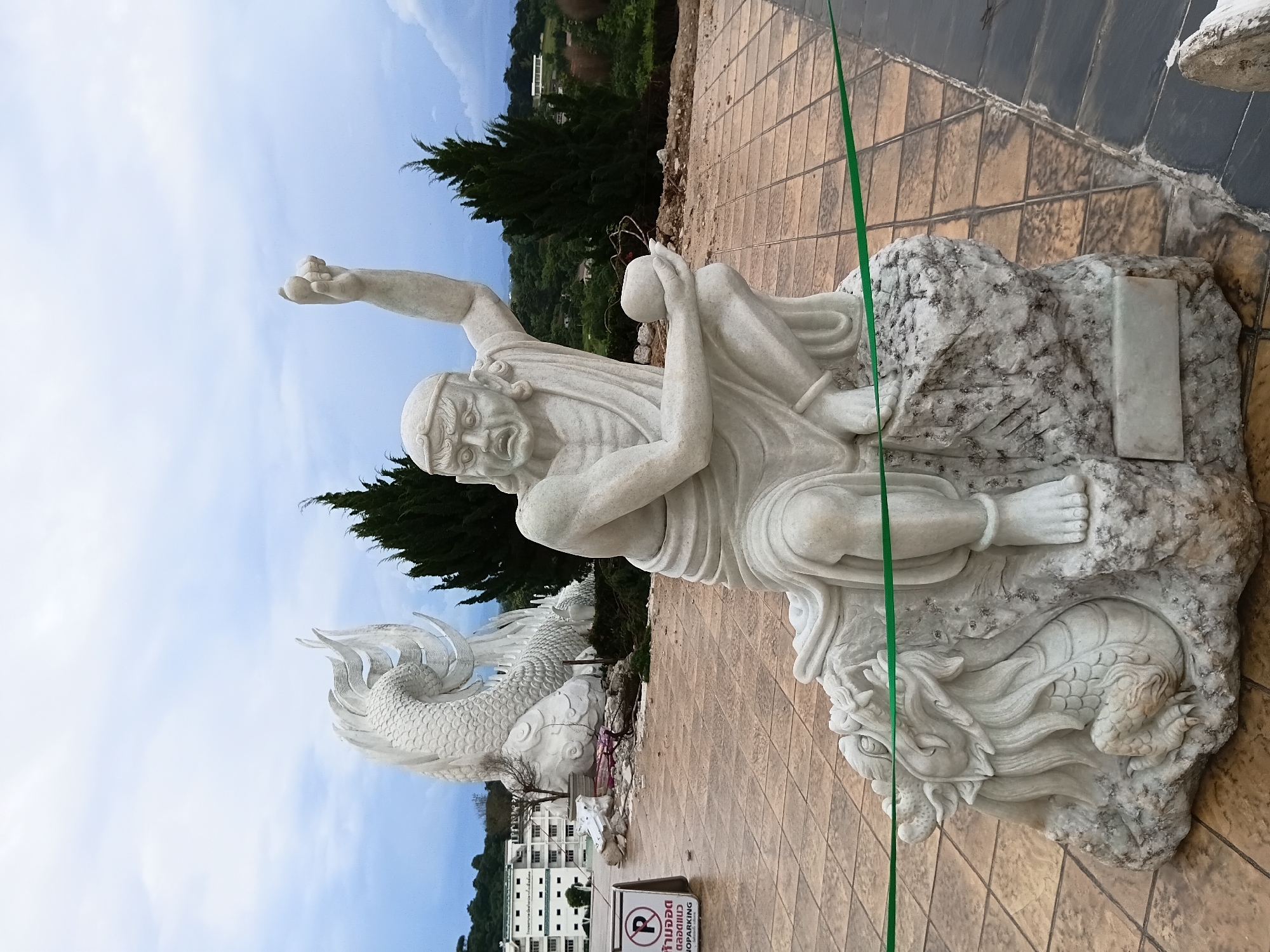 A white marble statue of a warrior figure is posed aggressively, squatting on a rocky base next to a dragon sculpture. The warrior has an intense expression with furrowed brows and an open mouth, one arm raised and clenched into a fist, while the other hand holds a round object. The statue is placed on a tiled plaza in front of a large, white dragon sculpture in the background. The environment appears to be outdoors with trees and buildings visible in the distance. A sign that reads "NO PARKING" is present on the left side of the image.