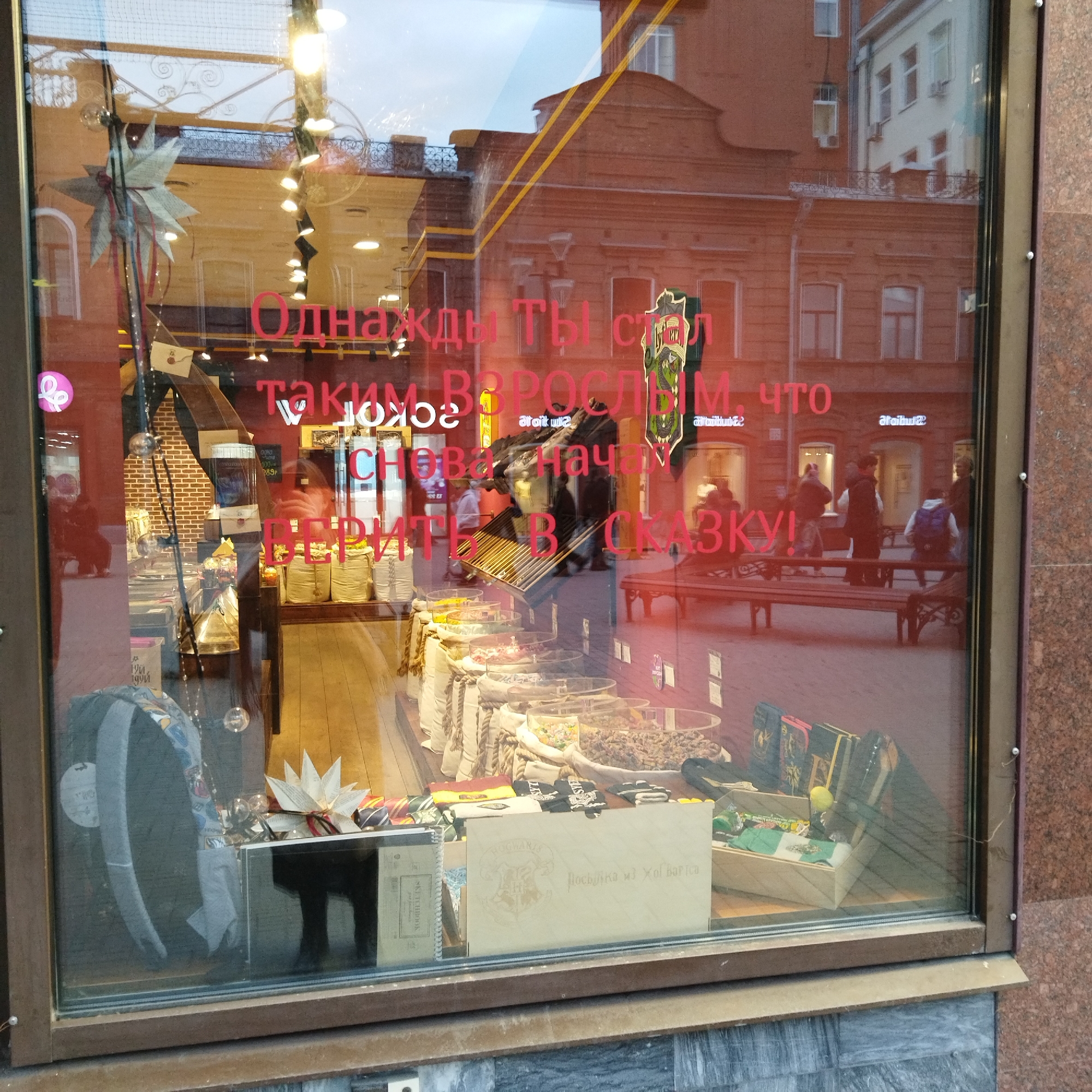 This image captures a storefront window in Yekaterinburg, Russia, displaying various items while also reflecting the street scene outside.
**Subjects & Scene:** The main subject is the shop window itself, with its interior contents and external reflections. Inside the shop, merchandise includes clear containers filled with what appears to be loose candy or dried fruits, displayed in front of white fabric bags with rope accents. There are also books, including a "SKETCHBOOK," and a wooden box with a crest and text. A decorative paper star hangs within the display. The exterior reflections show brick buildings, potentially an old-style edifice, with multiple windows and some ornate iron railings. Several indistinct figures of people are reflected, some walking and one person sitting on a bench in the mid-ground.
**Activity or Event:** The window showcases the shop's offerings, implying it is open for business. The reflected people suggest typical street activity with passersby.
**Notable Details:** The time of day appears to be daytime or early evening, as there is ambient light but no direct sun. The overall atmosphere is retail-oriented and somewhat whimsical, given the decorative elements and the messages. The presence of benches and brick buildings suggests an urban, possibly historic, street.
**Visible Text:** Prominently displayed in red letters on the inside of the window is a Russian phrase: "Однажды ты стал таким ВЗРОСЛЫМ, что снова начал ВЕРИТЬ В СКАЗКУ!" This translates to: "One day you became so ADULT that you started BELIEVING IN A FAIRY TALE again!" Another visible text is on a light wooden box inside the shop, which reads "Посылка из Хогвартса" ("Parcel from Hogwarts"), accompanied by a crest resembling the Hogwarts shield. A small price tag shows "Около 100-289р" (Approximately 100-289 rubles). Other partially visible text includes "SKETCHBOOK" and "ataittu@".