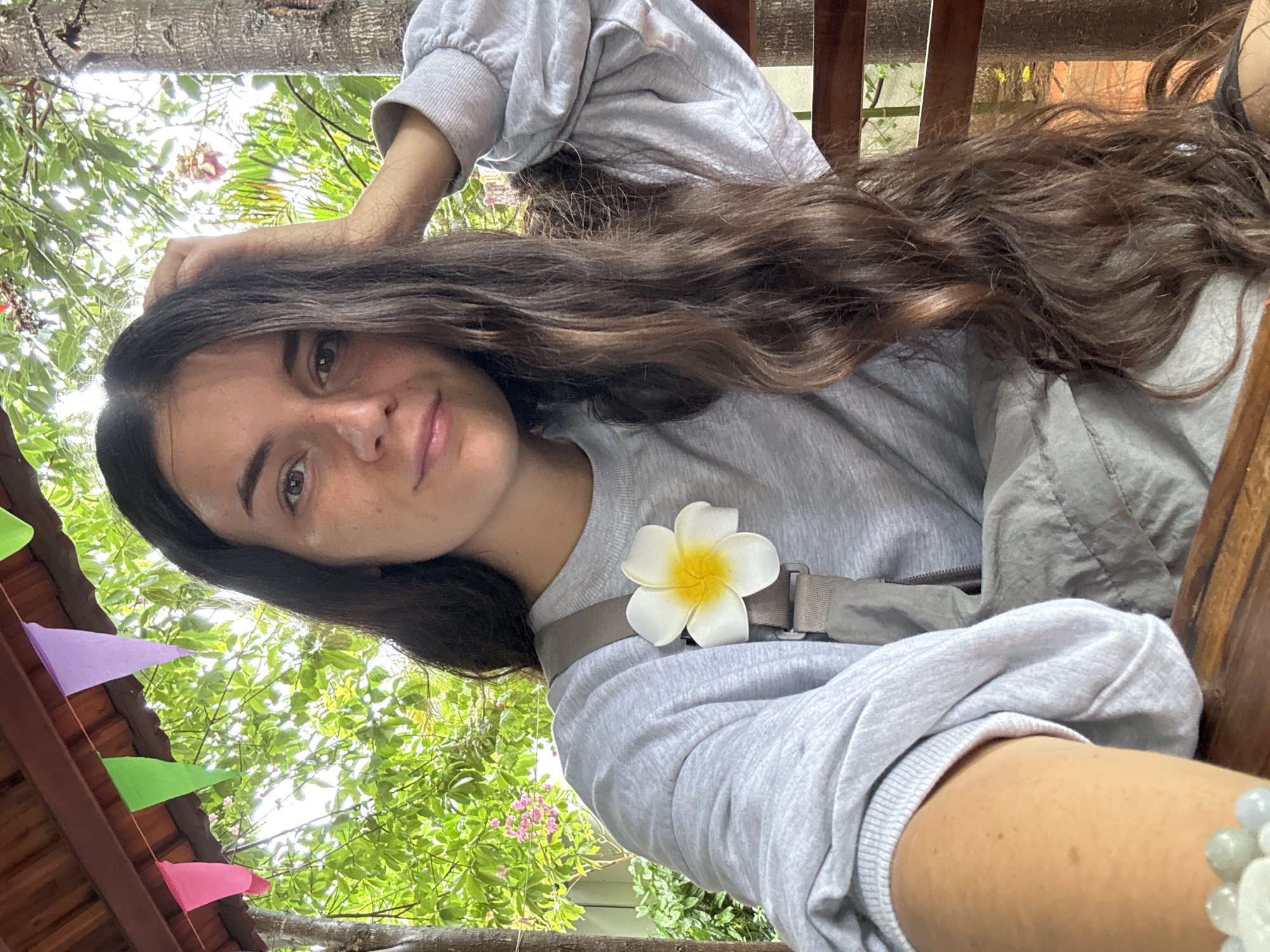 The photograph captures a young woman with long, brown wavy hair, fair skin, and visible freckles. She is looking directly at the camera with a slight smile. She is wearing a light gray long-sleeved sweatshirt and appears to be wearing a seatbelt over her shoulder, on which a white flower with a yellow center is affixed. Her right hand is resting on her head, and a bracelet is visible on her wrist.
The background shows a combination of natural and man-made elements. Lush green foliage is partially visible, suggesting an outdoor setting, possibly a garden or park-like area. There are some colorful triangular pennants attached to a wooden structure, which might be a roof or awning. The wooden structure suggests the scene is located in a rustic or outdoor eating area.
Based on the location specified, Mũi Né, Vietnam, the setting might be an outdoor cafe, restaurant, or a resort area, given the blend of nature and built elements. The presence of the seatbelt is unexpected, and it could be associated with a seat on a recreational vehicle or a swing chair in a tourist-oriented location.