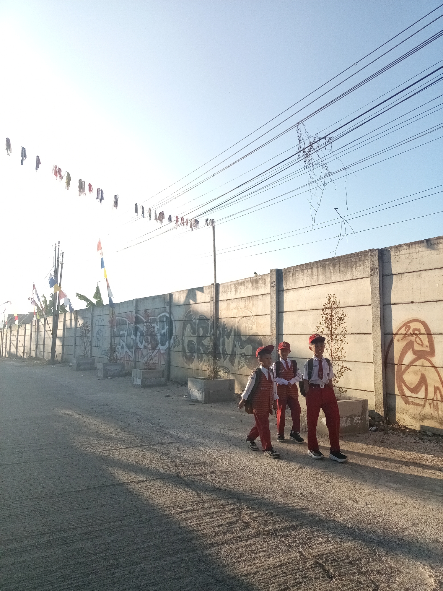 Three young boys in school uniforms, consisting of white shirts, red vests, red pants, and red caps, walk along a cracked asphalt road in Cigugur Girang, Indonesia. They are walking from left to right, with the boy on the left slightly ahead of the other two. The boys carry backpacks. Behind them is a tall, light-colored concrete wall with graffiti. In front of the wall are several rectangular concrete planters, some containing small trees. In the distance, a line of electrical wires stretches across the scene, with a few small flags or pennants strung along some of them. The sky is clear and blue, with the sun casting long shadows across the road.
