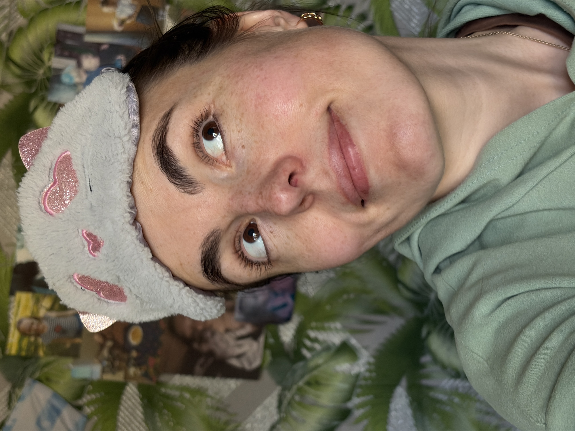 A woman is lying on a surface with a green leafy pattern. She is wearing a grey sleep mask with pink heart-shaped decorations and a light green top. Her head is turned to the side, and she is looking upwards. A gold earring is visible in her ear. Based on the provided information, the location is Nizhnii Vishur, Russia, though there are no visual cues confirming that location.