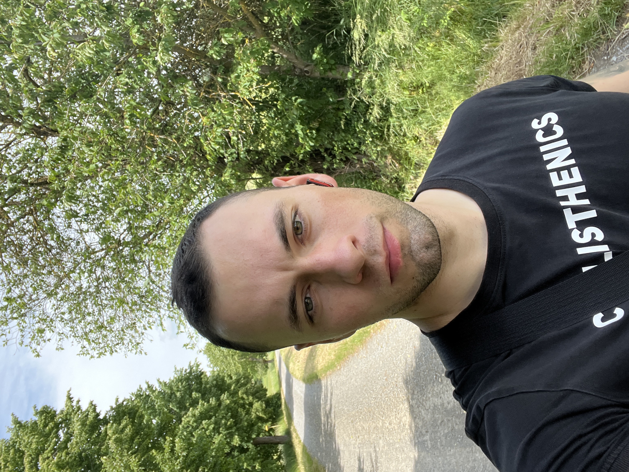 A man is depicted in a close-up self-portrait, with the image rotated 90 degrees clockwise. His head occupies the left side of the frame, facing towards the right, and he looks directly at the viewer with a neutral expression. He has short dark hair, light facial stubble, and green eyes. A red and black earbud is visible in his left ear.
He is wearing a black t-shirt with white capital letters that spell "STHENICS" vertically down the chest. A black strap, likely from a bag, is visible across his right shoulder.
The background, predominantly visible on the right side of the rotated frame, portrays a bright outdoor environment. Lush green trees with dense foliage and patches of green grass are prominent. A paved path, possibly asphalt or light-colored gravel, recedes into the distance, bordered by the abundant greenery. The sky, partially visible in the upper left section of the frame, appears bright and clear, indicating a daytime setting with fair weather. The scene suggests an activity or moment captured in a park or on a natural trail.