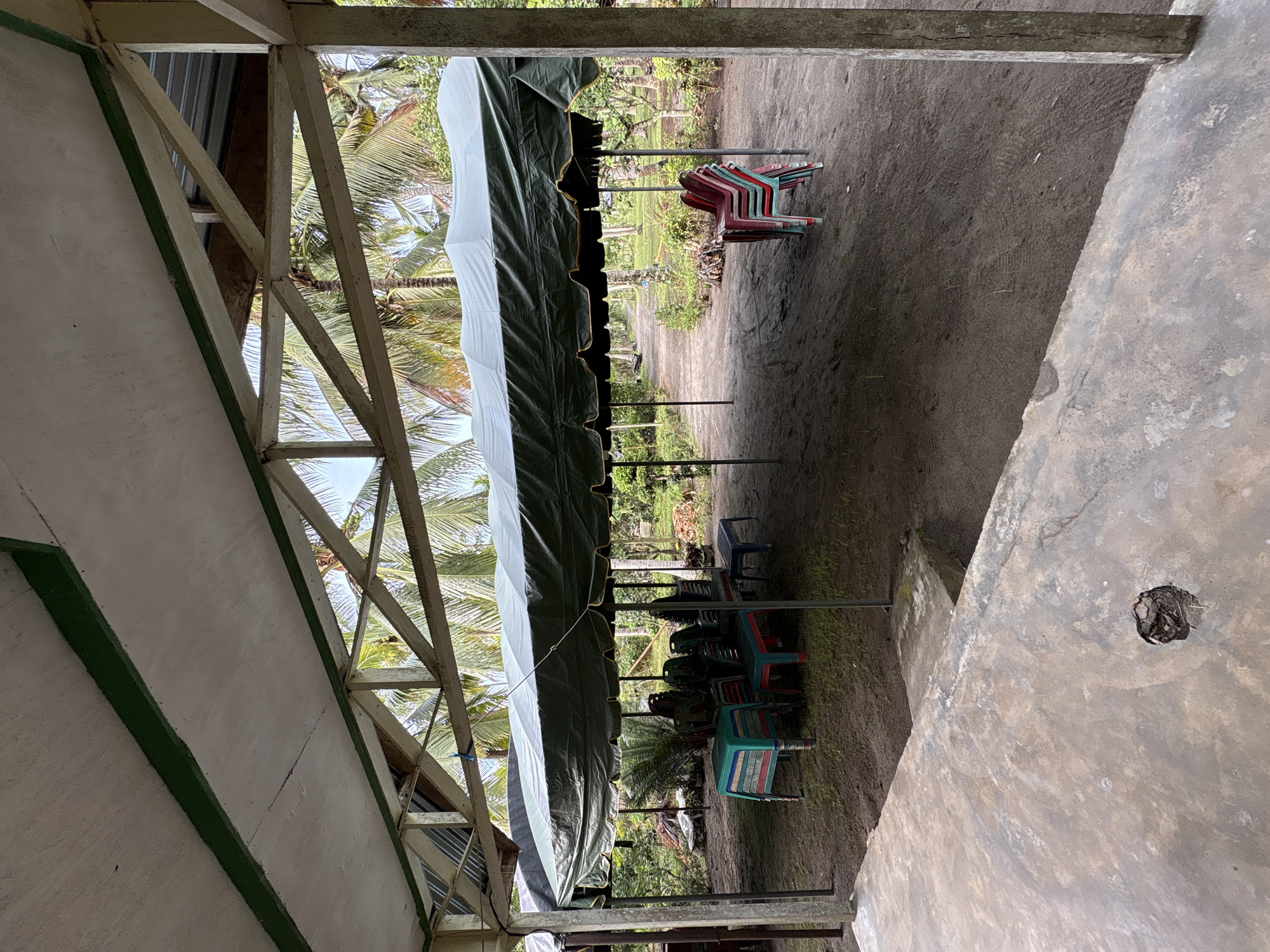 The image depicts an outdoor covered seating area, likely for an event, in Beunyot, Indonesia. A large dark green tent with a gold scalloped trim is supported by poles, creating shade over a sandy ground. Stacked plastic chairs in red, green, and white are visible under the tent. Some tables are also present, covered with colorful plastic. In the background, a path leads through a tropical landscape with numerous palm trees. The foreground features a concrete structure, possibly a patio or walkway, with a rough texture and a circular hole. The overall impression is of a simple, functional outdoor gathering space in a rural or semi-rural Indonesian setting.