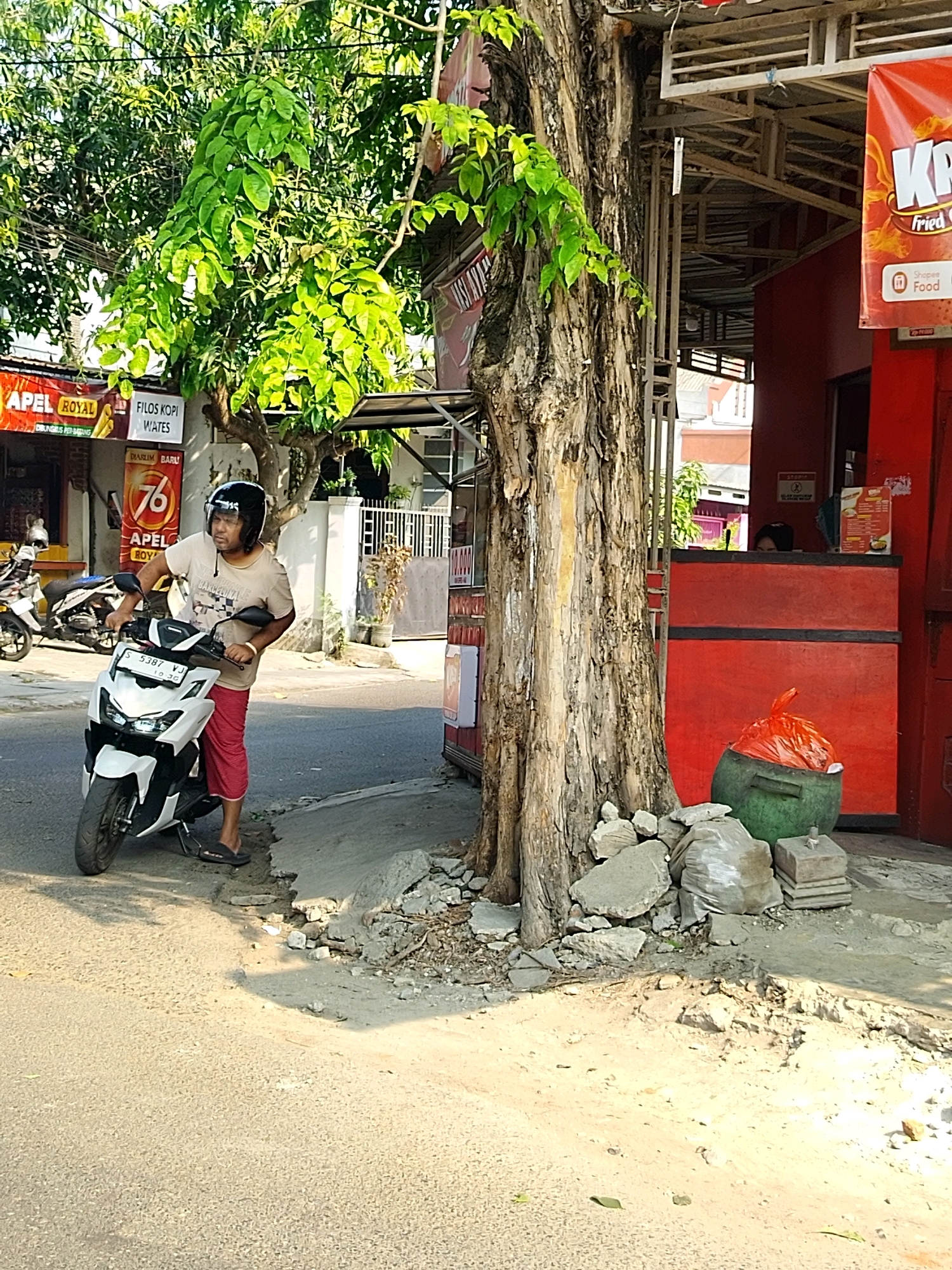 A man wearing a white t-shirt, red pants, a black helmet, and black sandals is standing next to a white and black motorcycle on a paved road in front of a building. The motorcycle has a license plate that reads "S 5387 VJ." Behind the man, there is a large tree with green leaves and a rough bark. To the right of the tree, there is a red building with a counter where two people are visible. A green trash bin with an orange plastic bag on top is placed in front of the building. Various signs are visible on the buildings, including one with "APEL ROYAL" and another with "FILOS KOPI WATES." The environment appears to be a street in the city of Mojokerto, Indonesia, possibly a commercial area with food vendors. The scene is lit by sunlight, creating shadows on the road and ground.