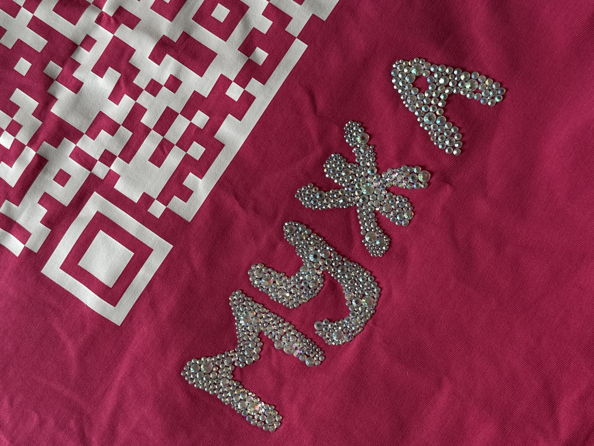 The image shows a pink fabric with a large white QR code design in the upper left corner. To the right, there are decorative shapes made of rhinestones: a "P", a flower, a "Y", and a "3." The setting appears to be indoors, with the fabric lying flat. There is no indication of the location being Izhevsk, Russia, or any people present.