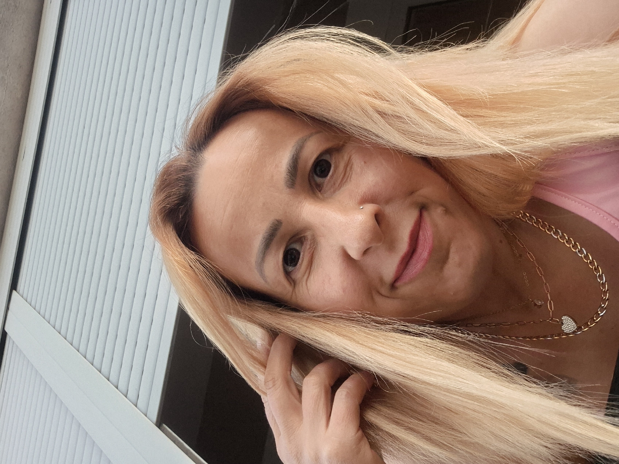 A selfie is taken from a low angle of a woman with blonde hair and light skin. She has a nose ring, earrings, and is wearing several necklaces. One necklace has a heart-shaped pendant. The woman is smiling and touching her hair with her right hand. She is wearing a pink top. In the background, a gray and white roller blind is visible. The setting appears to be indoors or on a covered patio.