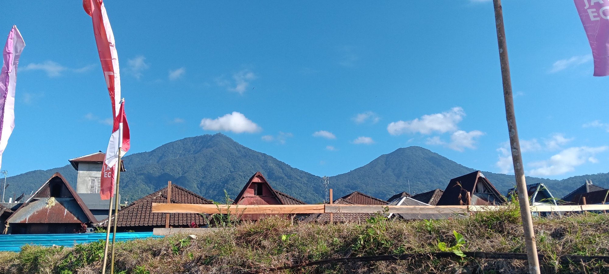 This is a wide-angle, eye-level outdoor shot taken in Penebel, Indonesia, on a clear day. The foreground is dominated by dry, brown grass and green foliage, with a wooden fence running horizontally across the middle ground. Behind the fence, several rooftops of houses are visible, some with rusty, corrugated metal and others with clay tiles. One house has a tall, square structure attached, possibly a watchtower or a part of the building. In the background, two large, verdant mountains rise against a bright blue sky dotted with a few white clouds. Two birds are visible flying in the sky. In the left of the frame, two flags are seen flying: one is a pale purple and white flag, and the other is a red and white Indonesian flag. A thick wooden pole is visible in the right foreground.