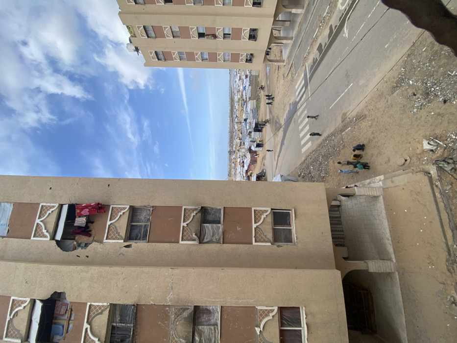 The image captures an elevated view of an urban landscape in Khan Yunis, Palestinian Territory, featuring residential buildings adjacent to a large displacement camp.
In the immediate foreground, a multi-story, light-brown residential building stands on the left. It exhibits significant structural damage, including large, irregular holes on its upper floors exposing interior areas. Clothes, including red garments, are visibly hanging to dry from one damaged window opening. Many windows on this building are covered with white or translucent material. The ground floor includes an arched, covered pedestrian area.
To the right, a similar multi-story building, also light-brown with decorative window frames, stands further back. It appears to have less visible exterior damage from this perspective and features water tanks on its roof.
Between these buildings and extending into the midground is an asphalt road with a white zebra crossing. The areas immediately surrounding the road are unpaved dirt and rubble. Approximately seven individuals, primarily children, are gathered on the dirt patch near the base of the left building. Two individuals are crossing the road via the zebra crossing, and other scattered figures are visible further down the road.
Beyond the road and unpaved areas, a vast, densely packed encampment stretches across the landscape towards the horizon. This encampment consists of numerous makeshift shelters and tents, predominantly white and light-colored, indicating a large-scale temporary settlement.
In the distant background, under a blue sky with scattered white clouds, a body of water, consistent with a coastline, is visible.