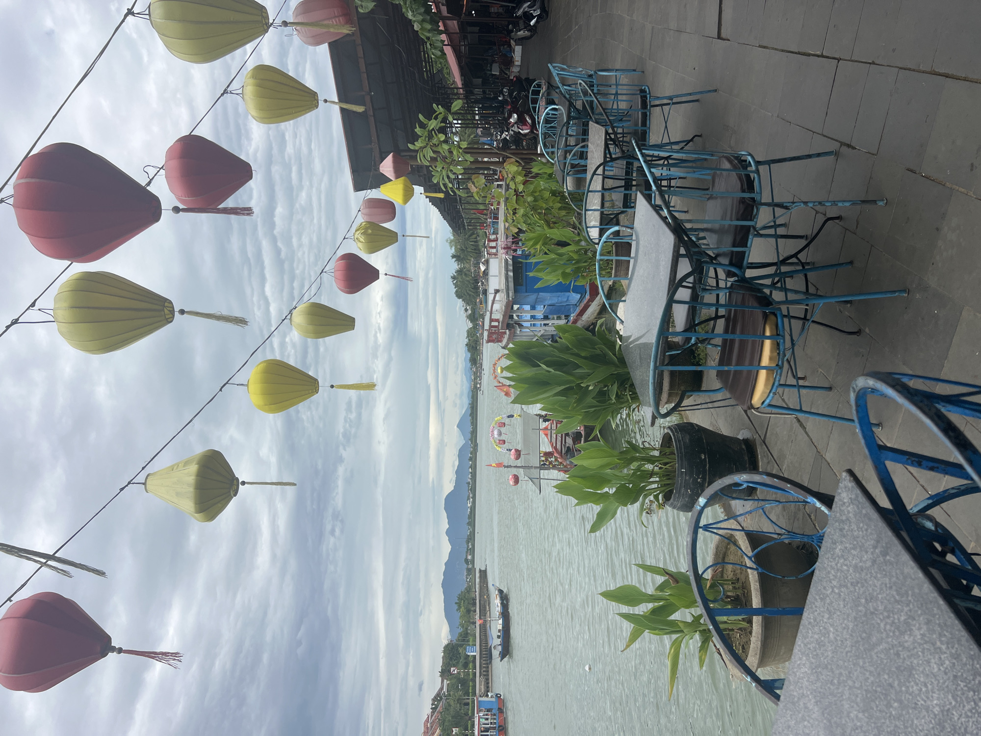 The image depicts a waterside promenade in Hoi An, Vietnam, with a river or canal on the left and a row of shops or restaurants on the right. The sky is overcast.
In the foreground, there are several outdoor tables and chairs. The tables have dark, speckled tops and are made of metal with a blue frame. The chairs are also made of metal with blue frames, and some have dark seat cushions. To the left of the tables, there are large potted plants with broad green leaves.
Beyond the tables and chairs, a body of water is visible, with several boats. One boat is a small, dark vessel with a sail. Another boat, further away, is larger and appears to be a passenger boat with a red and white structure. On the right side of the water, there is a long, covered walkway with shops or restaurants. The roofs are dark, and there are motorcycles parked in front of some of the establishments.
Hanging above the promenade, in the middle ground and background, are numerous colorful lanterns. These lanterns are of various shapes and colors, including red, yellow, and pink. They are strung across wires and create a festive atmosphere. In the distance, across the water, there are buildings and mountains.
The overall impression is a tranquil scene, possibly in the early morning or late afternoon, with the promise of a vibrant atmosphere when the shops and restaurants are fully open. The presence of the lanterns suggests a cultural or tourist-oriented area.
