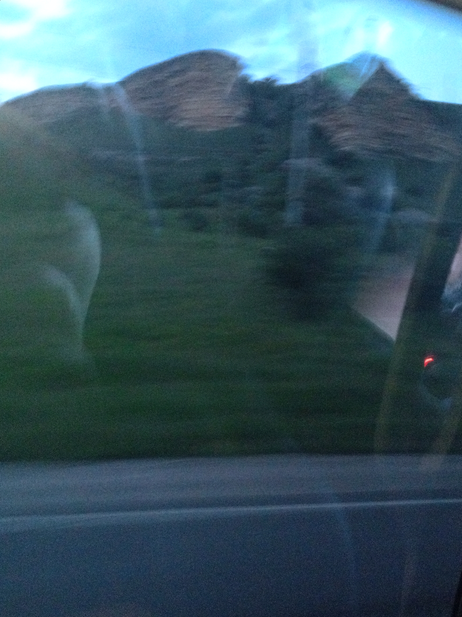 The image is a blurry, horizontal shot taken from inside a vehicle, looking out. The foreground shows a dark blue surface, likely the interior of the car. The middle ground is a blurred green expanse, suggesting grass or foliage. In the background, a large, dark, mountainous or hilly landscape is visible, with a bright, hazy sky above. The blurriness suggests movement, likely the car in motion. There is no discernible indication of the specific location within Ali-Berdukovskiy, Russia.