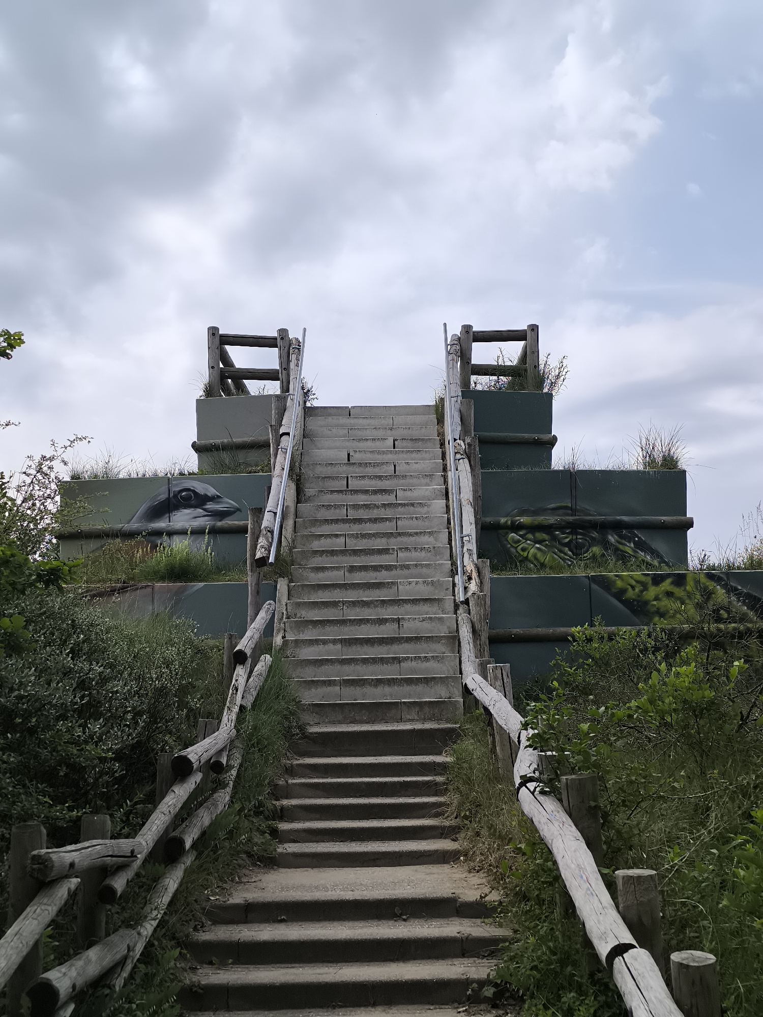 A full shot depicts a set of concrete stairs leading up to an elevated structure in Zandvoort, Netherlands. The stairs are flanked by weathered wooden railings on either side. The elevated structure consists of tiered, green-painted concrete blocks adorned with wildlife murals – a bird's head on the lower tier and a reptile on the upper tier. Patches of grass sprout from the top of the blocks. Lush greenery and vegetation border the stairs, adding to the natural setting. The sky above is overcast with a mix of white and gray clouds.