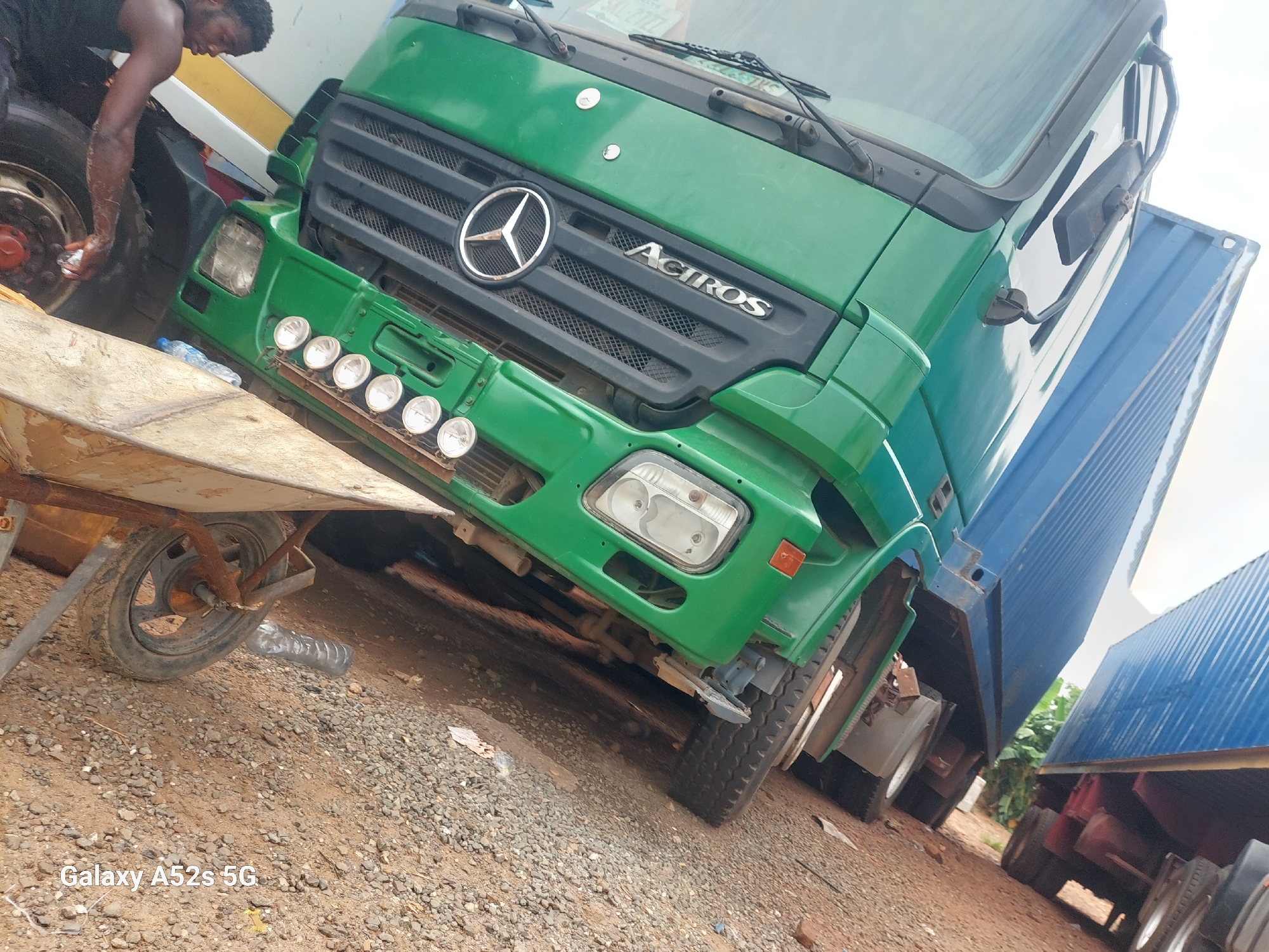 In Awkuzu, Nigeria, a green Mercedes-Benz Actros truck is parked on a dirt and gravel surface, with a large blue shipping container attached. The truck's front grille features the Mercedes-Benz star logo and the "Actros" model name. Below the grille, there are several round auxiliary lights. A man in a black tank top is visible to the left of the truck, appearing to be attending to the front tire and holding a clear plastic bottle. A wheelbarrow with a single wheel is positioned in the foreground, to the left of the truck, resting on the ground. The environment appears to be outdoors with a bright, overcast sky. Another truck, partially visible, is parked behind the blue shipping container.