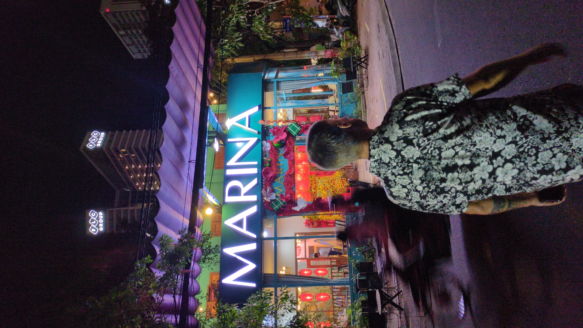 This image depicts a nighttime urban scene in South Quy Nhon Ward, Vietnam. The primary subject is a male individual, viewed from behind, with short, graying hair, wearing a short-sleeved black shirt with a white floral pattern. A tattoo is visible on his left arm, and his right hand is held low, possibly gripping an object. He stands on a paved road, facing towards a brightly lit building in the midground.
The building in the midground features a prominent white neon sign displaying "MARINA" in capital letters. Its exterior is turquoise, with large glass windows revealing an interior decorated with red lanterns, numerous yellow floral arrangements (likely for Tet/Lunar New Year), red paper decorations, and square green/red motifs resembling traditional Vietnamese glutinous rice cakes. Inside, a person in a light-colored shirt is visible standing near a doorway. Several dark outdoor tables and chairs are arranged on the sidewalk in front of the "MARINA" establishment. A blurred red streak of light and motion, consistent with a passing motorcycle or scooter, is visible to the left of the primary subject.
Above the "MARINA" building, a white, pleated canopy or awning is illuminated with purple light. In the distant background, against a dark night sky, two tall, multi-story buildings are visible. Each building's roof features a lit sign reading "FLC GROUP". Additional urban structures, streetlights, and green foliage are visible on both sides of the street, with parked motorcycles faintly discernible on the right side near trees. The scene is illuminated by a combination of streetlights and the numerous lights emanating from the buildings and decorations.