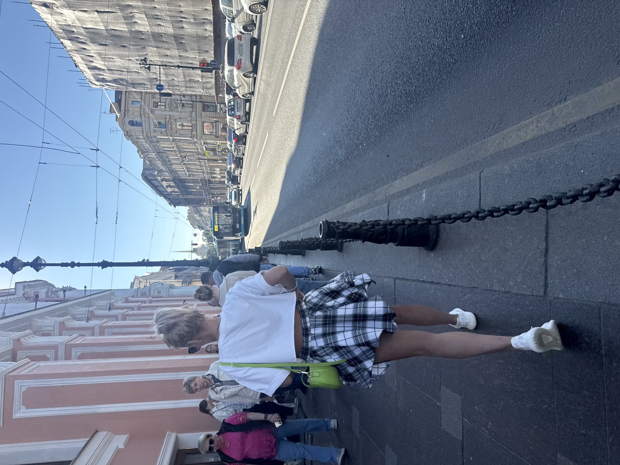 The image shows a street scene in Saint Petersburg, Russia, with a clear blue sky indicating daytime. Several people are walking on a sidewalk next to a street with traffic. The architecture of the buildings suggests an urban environment.
In the foreground, a woman wearing a white cropped t-shirt and a black and white plaid skirt is walking away from the camera. She has a bright green crossbody bag. Her legs are visible, and she is wearing white sneakers. Other people are visible in the background, also walking on the sidewalk. Some are wearing casual clothing.
The street is lined with cars, and there are overhead wires, likely for public transportation. A large building with ornate architecture is visible on the left side of the image. In the distance, more buildings and streetlights can be seen. The overall mood appears to be casual and everyday.
There are no overt emotional expressions visible, but the bright lighting and clear sky suggest pleasant weather. The presence of multiple people walking and the general urban setting indicate a public space in use. The scene appears to be a typical day in the city. Some road signs are visible in the distance, but the text is not legible.