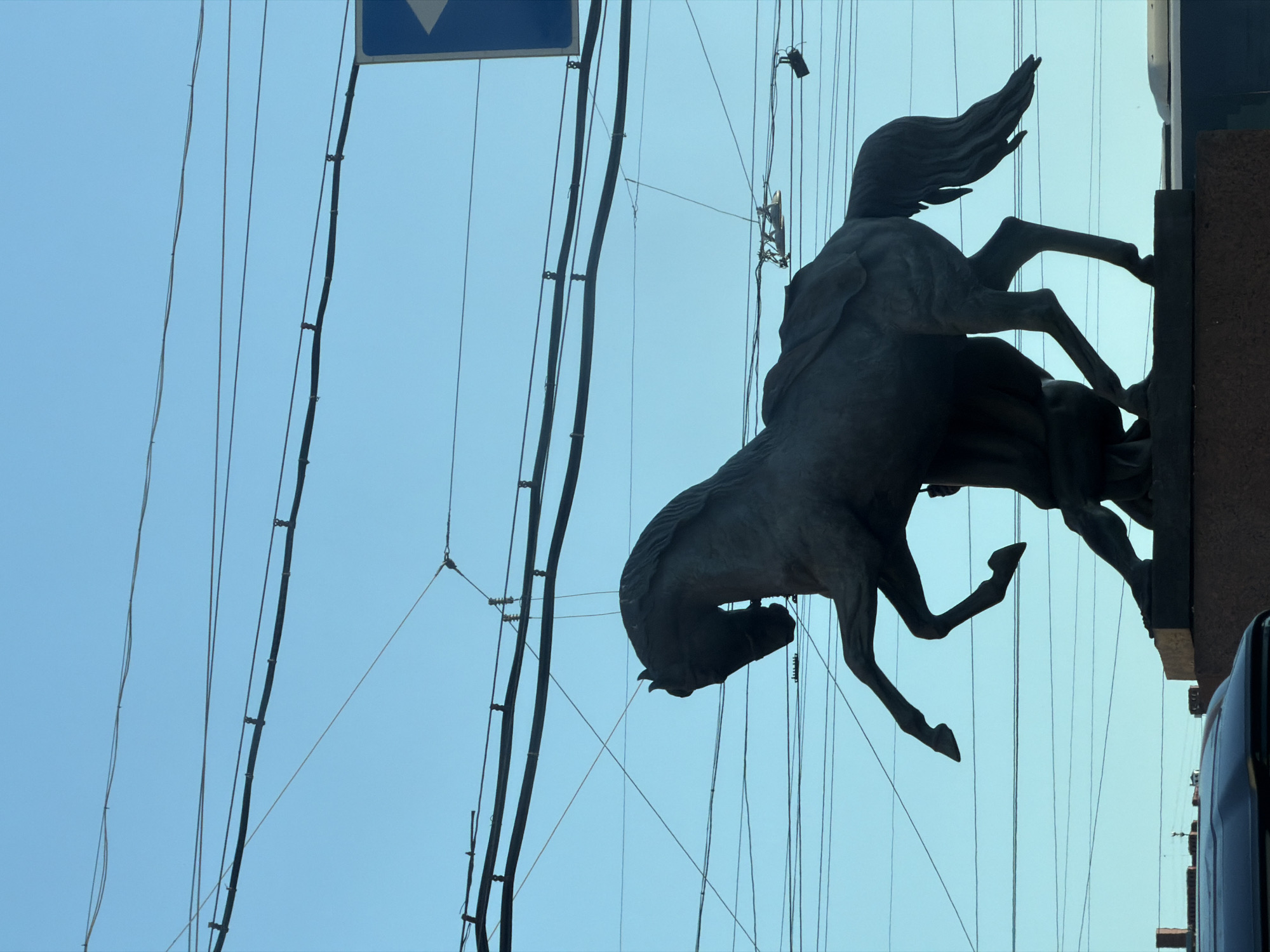 The image captures a silhouette of a bronze equestrian statue against a clear blue sky crisscrossed by numerous overhead wires. The statue depicts a horse rearing up on its hind legs, with a human figure beneath it, appearing to be restraining or controlling the animal. The horse is muscular and detailed, with its mane flowing and tail extended. The human figure is similarly rendered, with visible musculature and clothing. The statue is positioned on a plinth, which is partially visible at the bottom of the frame. In the background, a traffic sign with a blue background and a white arrow is visible. The overhead wires suggest an urban setting, likely a street with a tram or trolleybus system. The style of the statue and the presence of overhead wires are characteristic of Saint Petersburg, Russia.