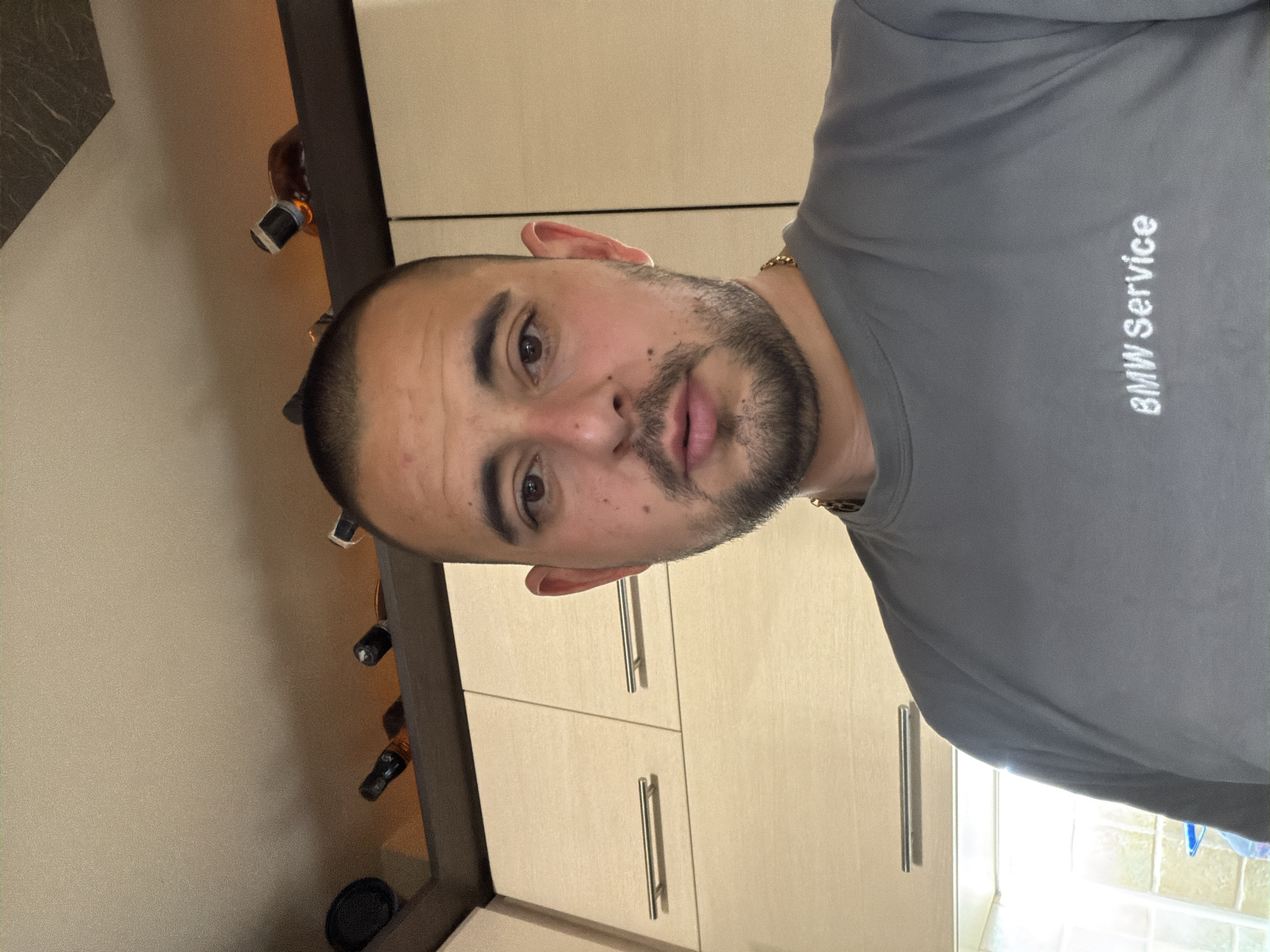 The image shows a man in a gray "BMW Service" shirt indoors, possibly in a kitchen based on the presence of cabinets and a wine rack. The man is facing the camera, and appears to be taking a selfie. There are no obvious details that link the scene to Burgas, Bulgaria.