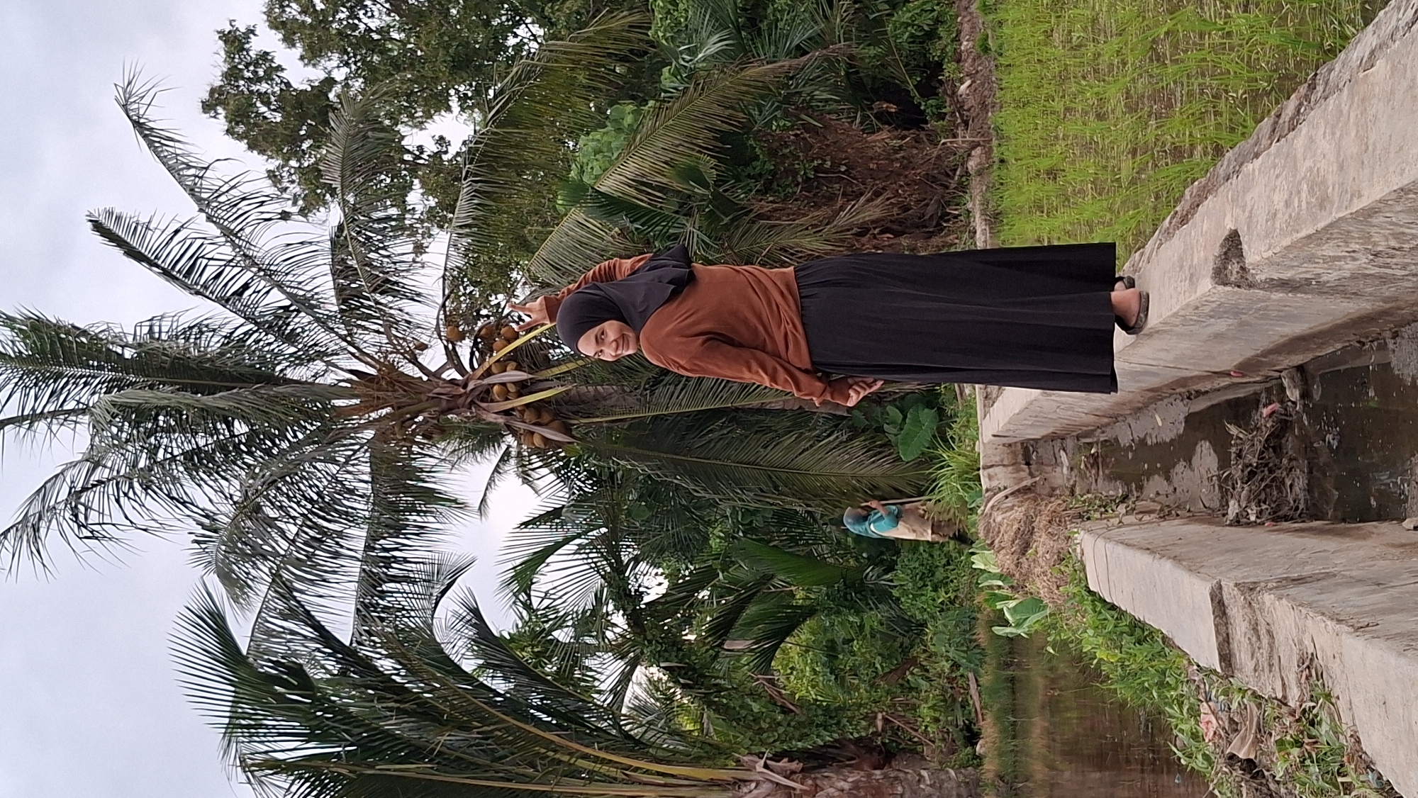 In Kutaglumpang, Indonesia, a woman wearing a black hijab and a brown long-sleeved top stands on a concrete pathway. She is looking back over her left shoulder, smiling, with her right arm raised in a V-sign gesture. Behind her, a coconut tree is laden with coconuts. Further in the background, another person, possibly a farmer, is visible tending to the fields. The environment consists of lush green vegetation, including palm trees and a rice paddy, with a narrow water channel running alongside the pathway. The sky is overcast.