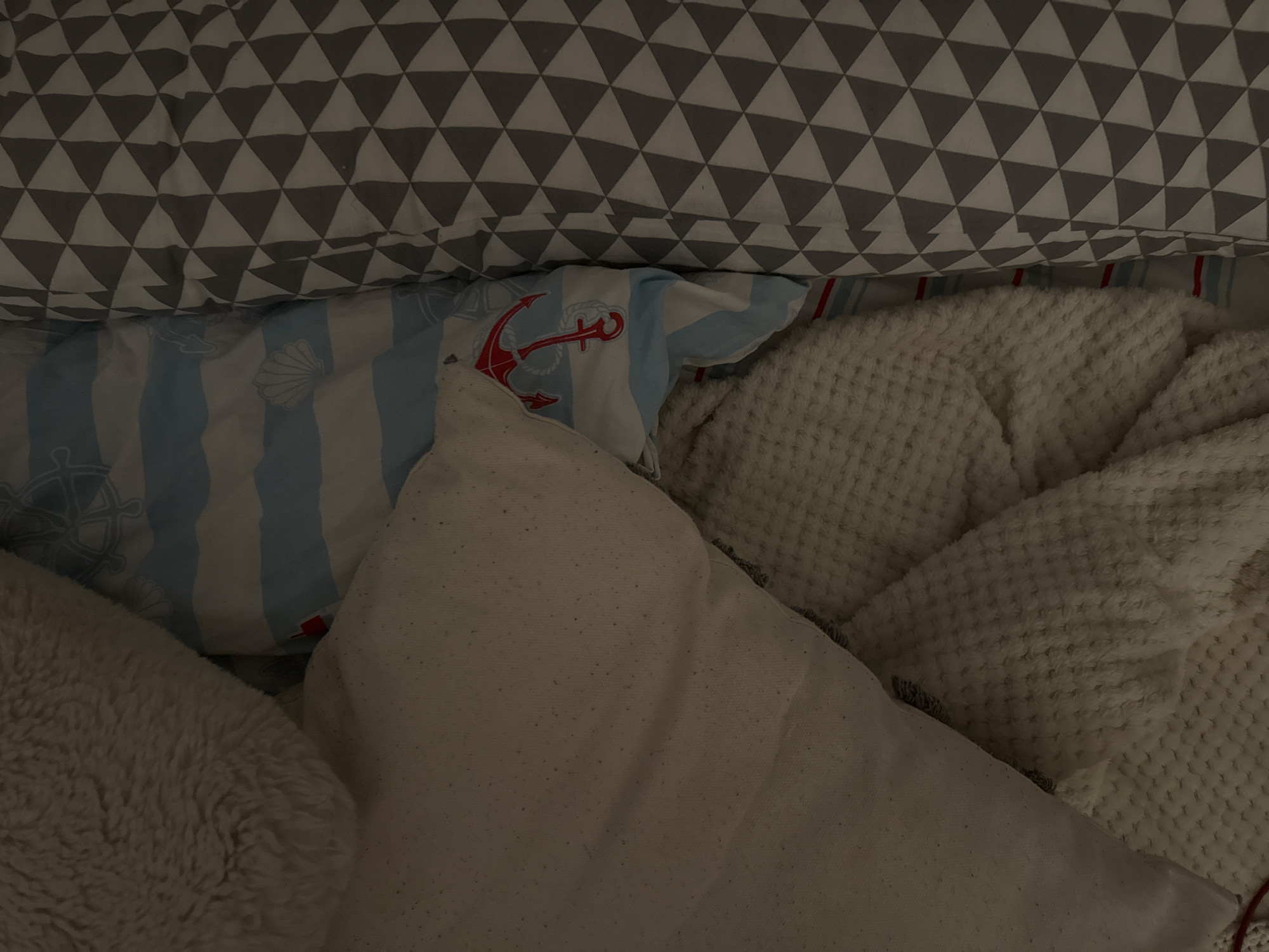 A close-up, overhead shot shows a pile of bedding in dim lighting. The bedding consists of several layers with different patterns. On the left, a soft, fluffy, beige blanket is visible. Next to it, a light-colored pillow with a textured surface is partially covered by the blanket. Beneath the pillow, a blue and white striped fabric with a red anchor pattern is peeking out. To the right of the striped fabric, a gray and white patterned blanket with repeating triangles is laid out. The bedding appears to be on a bed. The image is taken in Izobilnyy, Russia.