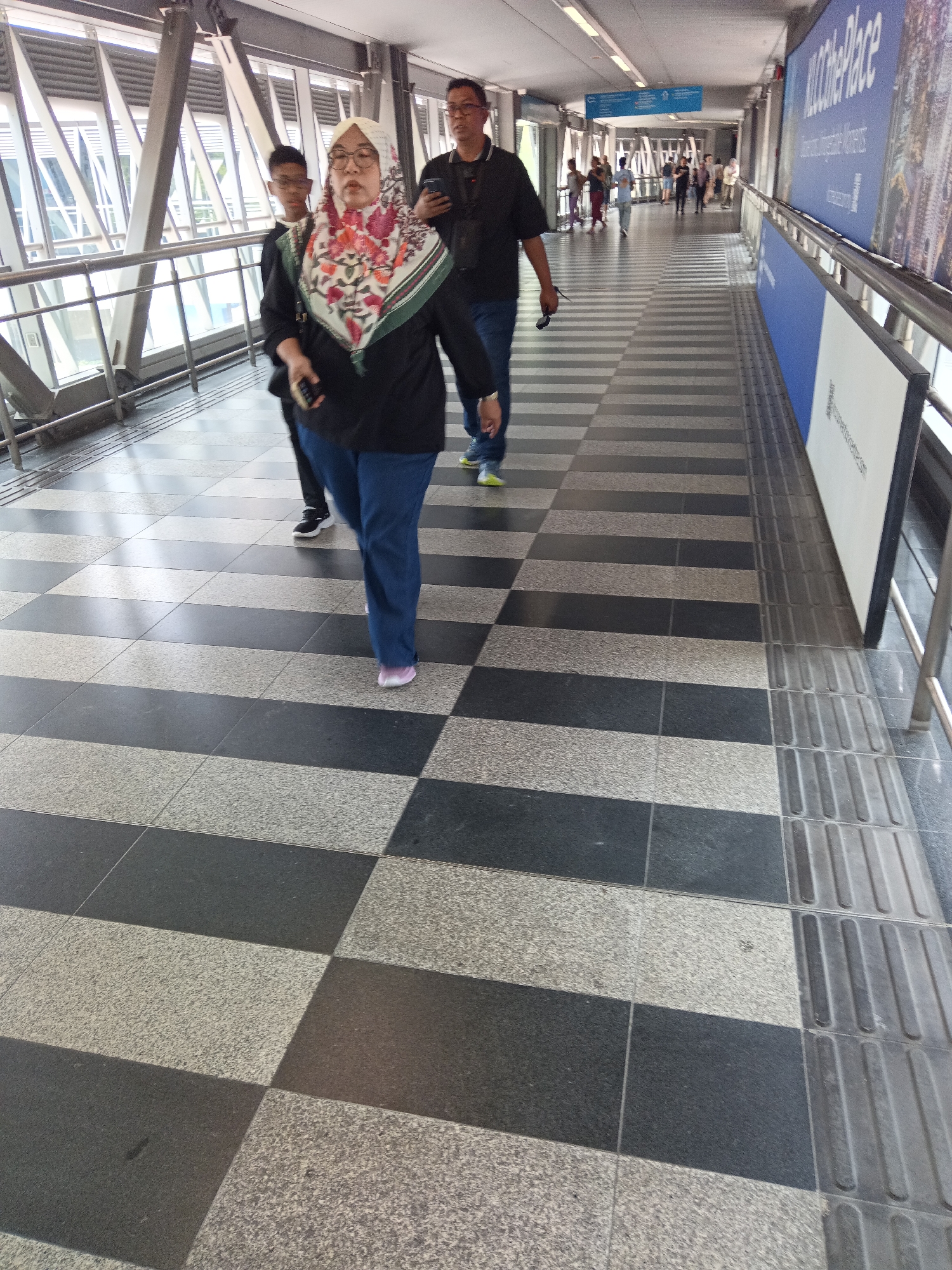 The image shows a group of people walking on a pedestrian bridge in Kuala Lumpur, Malaysia. The bridge has a checkered floor pattern of light gray and dark gray tiles. On the right side of the bridge, there is a large blue and white advertisement board. In the foreground, a woman in a hijab and blue pants is walking away from the camera. She is carrying a black bag in her left hand. To her left, a young boy in a black shirt and dark pants is also walking away from the camera. Behind them, a man in a black shirt and blue jeans is walking towards the camera, holding a mobile phone in his right hand. In the background, several other people are walking on the bridge. The bridge is enclosed by glass panels and metal railings. The lighting appears to be artificial, suggesting an indoor or covered outdoor space.