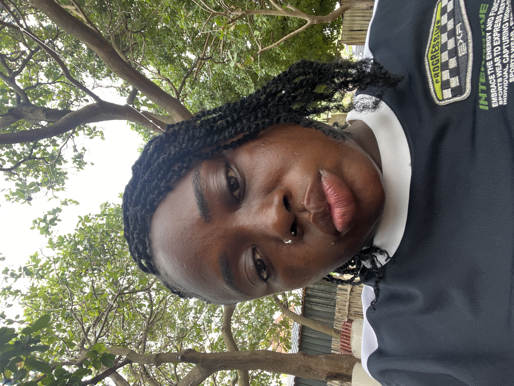 A close-up, front-facing selfie shows a young Black woman with braided hair and a nose ring, with lush green trees and a cloudy sky in the background. She is wearing a black t-shirt with a white collar and a patch on the left side of her chest that reads "PROGRESSIVE" and "EXR" within a checkered pattern. The text below the patch reads "INTERACTIVE EMBRACE THE ENERGY AND FUNCTION SPORTS WEAR TO EXPRESS REFLECT INDIVIDUAL. CAPTURING HIGH-END SPORTS WEAR-EXR". The background reveals a residential setting with a corrugated metal fence and bamboo fencing. The woman's facial expression is neutral, with her lips slightly pursed. The overall impression is that of a casual outdoor portrait. This image was taken in Abuja, Nigeria.