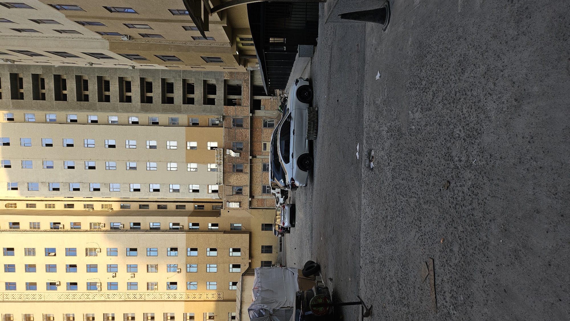This is a full, eye-level, exterior shot of a multi-story apartment complex in Dushanbe, Tajikistan. The buildings are tall and modern, with many windows and balconies. The complex appears to be under construction in some areas, with exposed brickwork and unfinished concrete visible. There is a parking lot in front of the buildings, with several cars parked. The ground is paved with asphalt, and there is some debris scattered around. The sky is overcast.