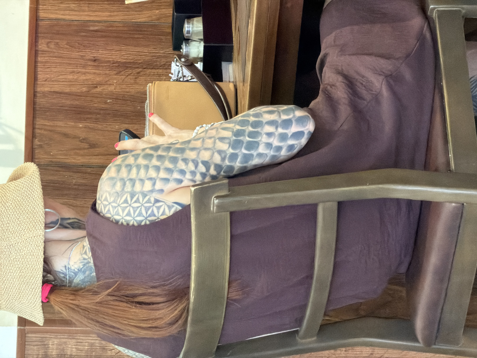 A woman with extensive tattoos on her arms and back, wearing a straw hat and a purple dress, is seated on a chair at a wooden table. Her hair is tied back with a pink scrunchie. She is looking at a smartphone in her hand. On the table in front of her are a tan wallet, a black box, and two small glass containers. The background consists of wooden paneling. This scene appears to be indoors, possibly a restaurant or cafe, located in Nha Trang Ward, Vietnam.