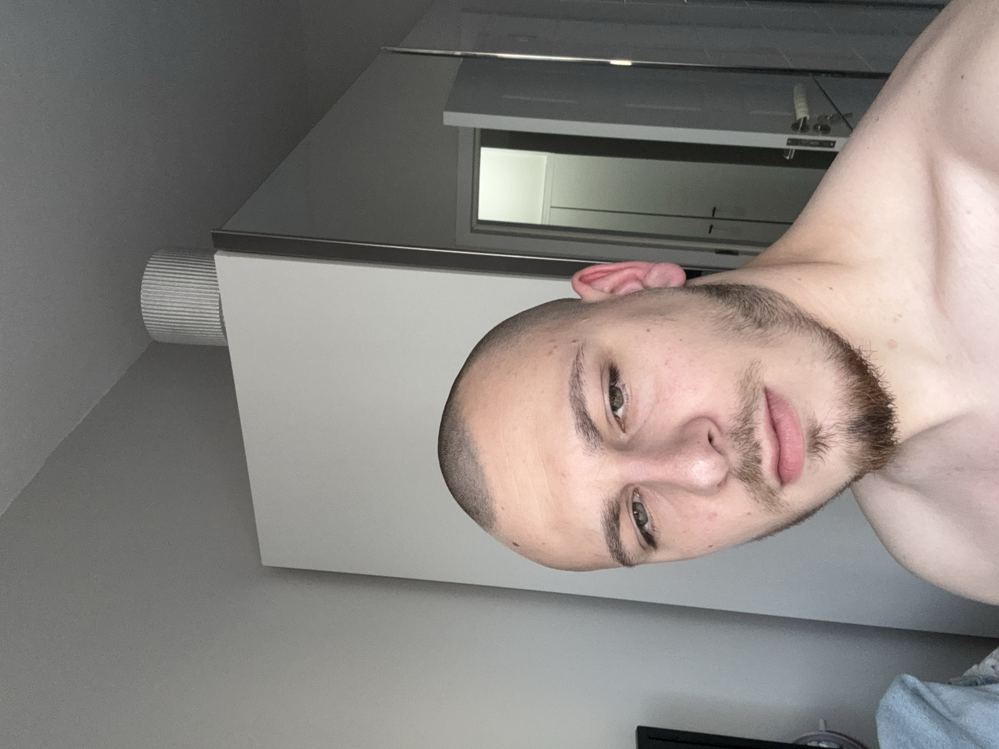 A vertically oriented photograph depicts a Caucasian male with a shaved head and a beard. He is positioned in the foreground, facing the camera with a slight downward angle. His skin tone appears fair, and his eyes are open and looking towards the camera. The background reveals an interior setting, featuring a wall painted in a light shade of gray, a ventilation shaft, and a door. The location is likely an indoor space, possibly a bathroom or bedroom, but there is no evidence to pinpoint the location to Stara Zagora, Bulgaria. No actions or interactions are depicted.
