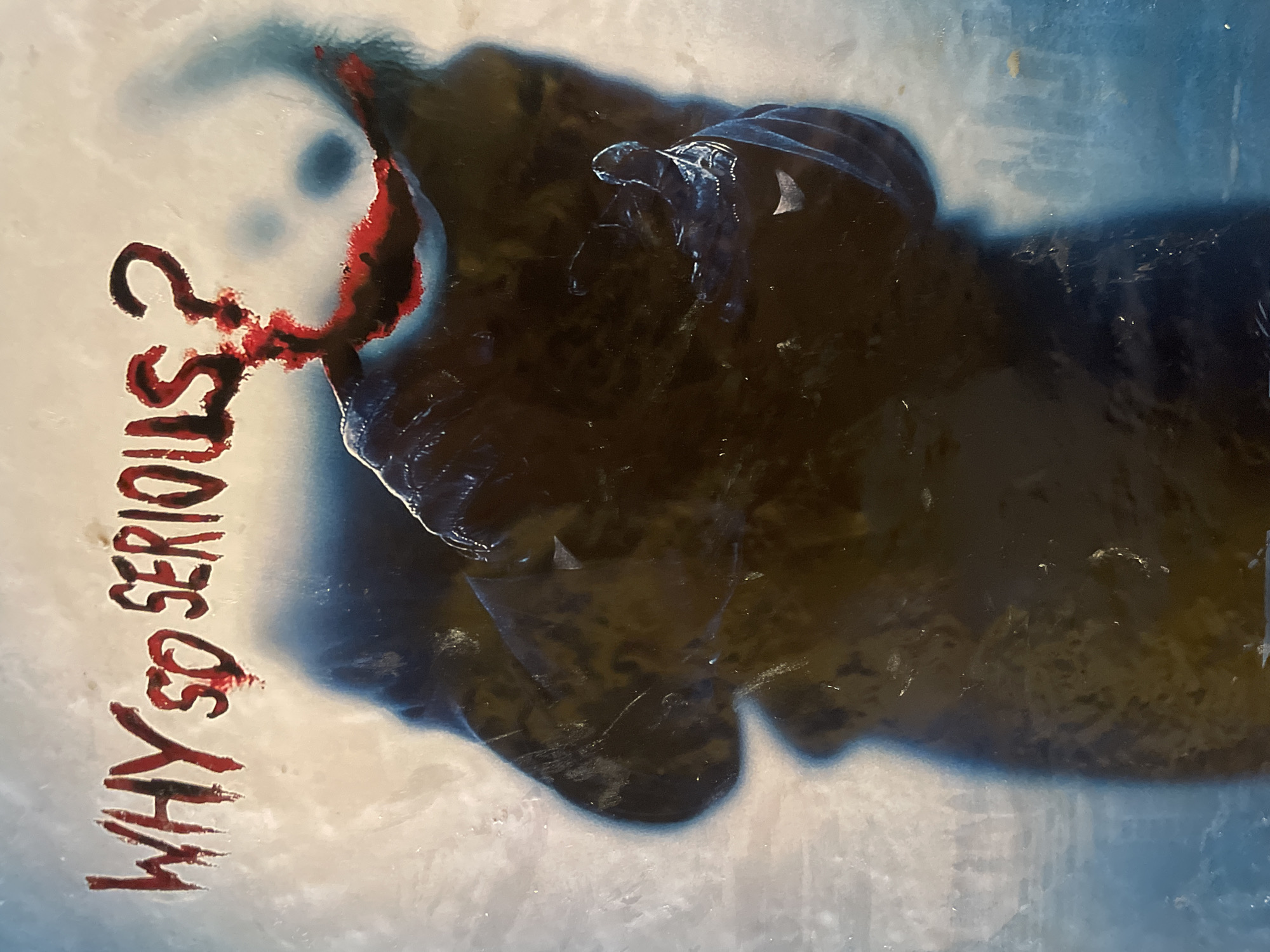 The image shows a close-up of a poster or artwork featuring the text "WHY SO SERIOUS?". The text is rendered in a dripping, blood-red font. Behind the text, a dark, shadowy figure is depicted, with a blurred suggestion of a smiling face painted in white and red. The background is a textured, abstract blend of light blue and white, with hints of a darker blue shadow cast by the figure. No specific location in Vagator, India, is visible or indicated in the image.
