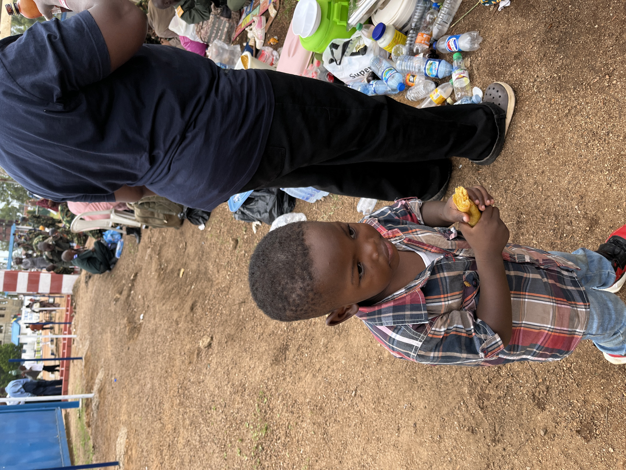 In Barakin Lerawi, Nigeria, a young boy in a plaid shirt and jeans stands on a dirt ground, holding a piece of food. In the background, adults in various attire, some in military uniforms, are visible near a structure with red and white striped columns and a blue wall. Several water bottles and plastic containers are scattered on the ground.