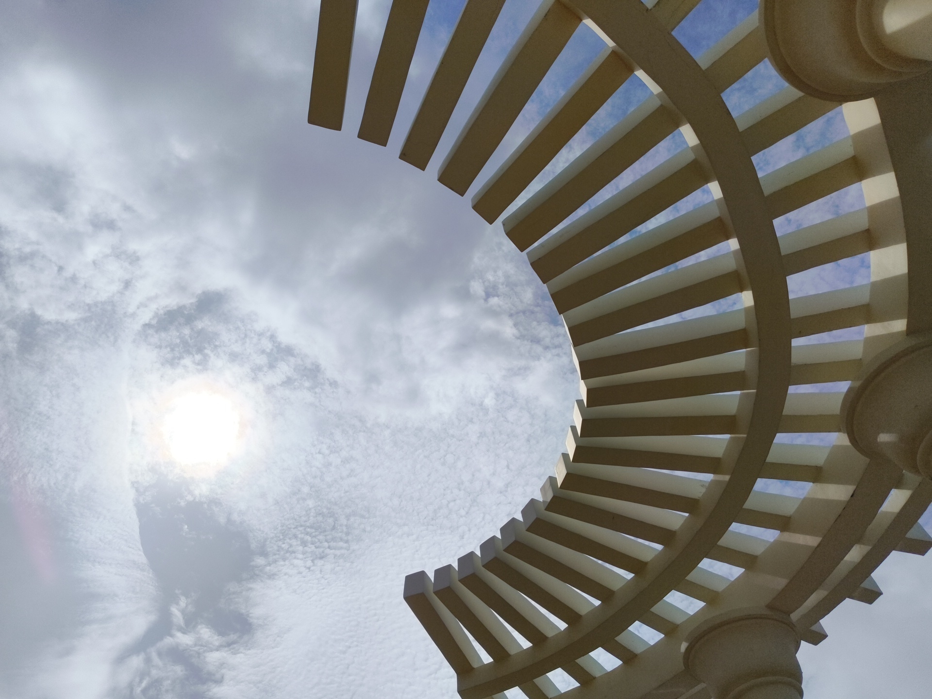 An upward-facing, low-angle shot captures a circular pergola structure against a cloudy sky. The pergola, made of cream-colored beams and columns, forms a sweeping arc across the frame. Sunlight shines brightly through the clouds on the left side of the image, casting a lens flare. The sky is a pale blue with scattered white clouds. The scene is located in Phú Quốc, Vietnam, a popular tourist destination known for its beaches and resorts.