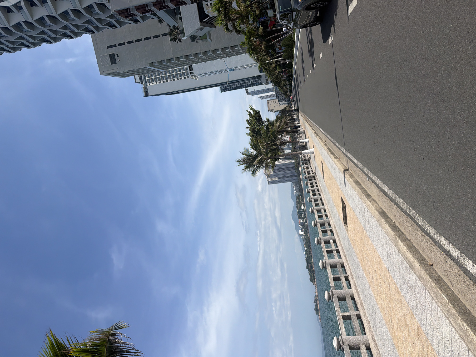 The image shows a sunlit, wide, paved road running parallel to a coastline in Bac Nha Trang Ward, Vietnam. Tall, modern buildings with balconies line the right side of the road, interspersed with palm trees and some smaller trees. A dark-colored SUV is parked on the right side of the road. A sidewalk, paved with light-colored stones and patterned sections of yellow and gray, runs alongside the road, separated by a concrete curb. Beyond the sidewalk is a stone promenade with a railing overlooking a body of water, likely the sea. The water is a deep blue-green, and the horizon shows a distant coastline with hills and buildings. The sky is a clear blue with scattered, wispy white clouds, and the bright sunlight casts shadows. A single palm frond is visible in the upper left corner, suggesting lush vegetation.