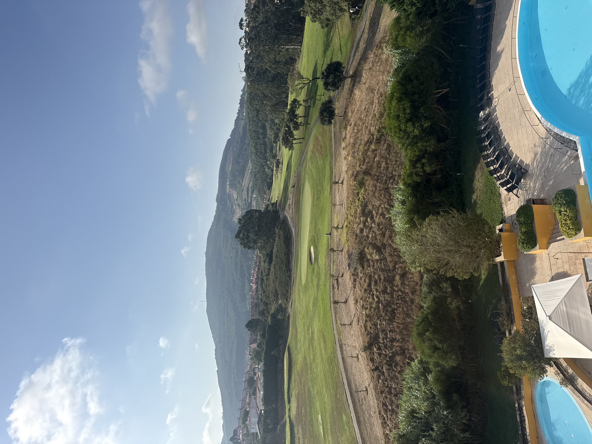 The image depicts a panoramic view of a golf course and surrounding landscape under a clear blue sky with scattered white clouds. In the foreground, a bright blue swimming pool with lounge chairs and a white canopy is visible, suggesting a resort or hotel setting. The golf course features well-maintained green fairways, sand traps, and scattered trees, with a hilly, tree-covered terrain in the background. A cluster of residential buildings with terracotta roofs can be seen nestled on the hillside. The overall scene is serene and suggests a leisure or vacation destination. This scene is located in the city of Torres Vedras, Portugal.